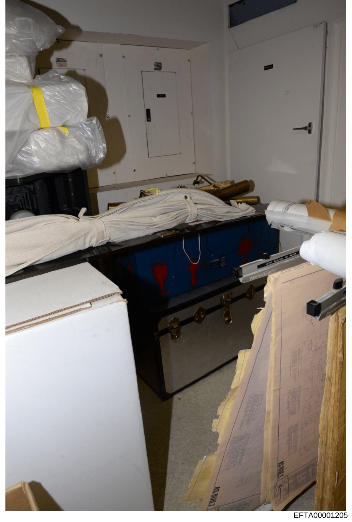 This is a photograph of an evidence storage or property room containing seized items wrapped in protective plastic and stored on shelving. The image shows a blue/black trunk or container in the center, large plastic-wrapped bundles (some marked with 