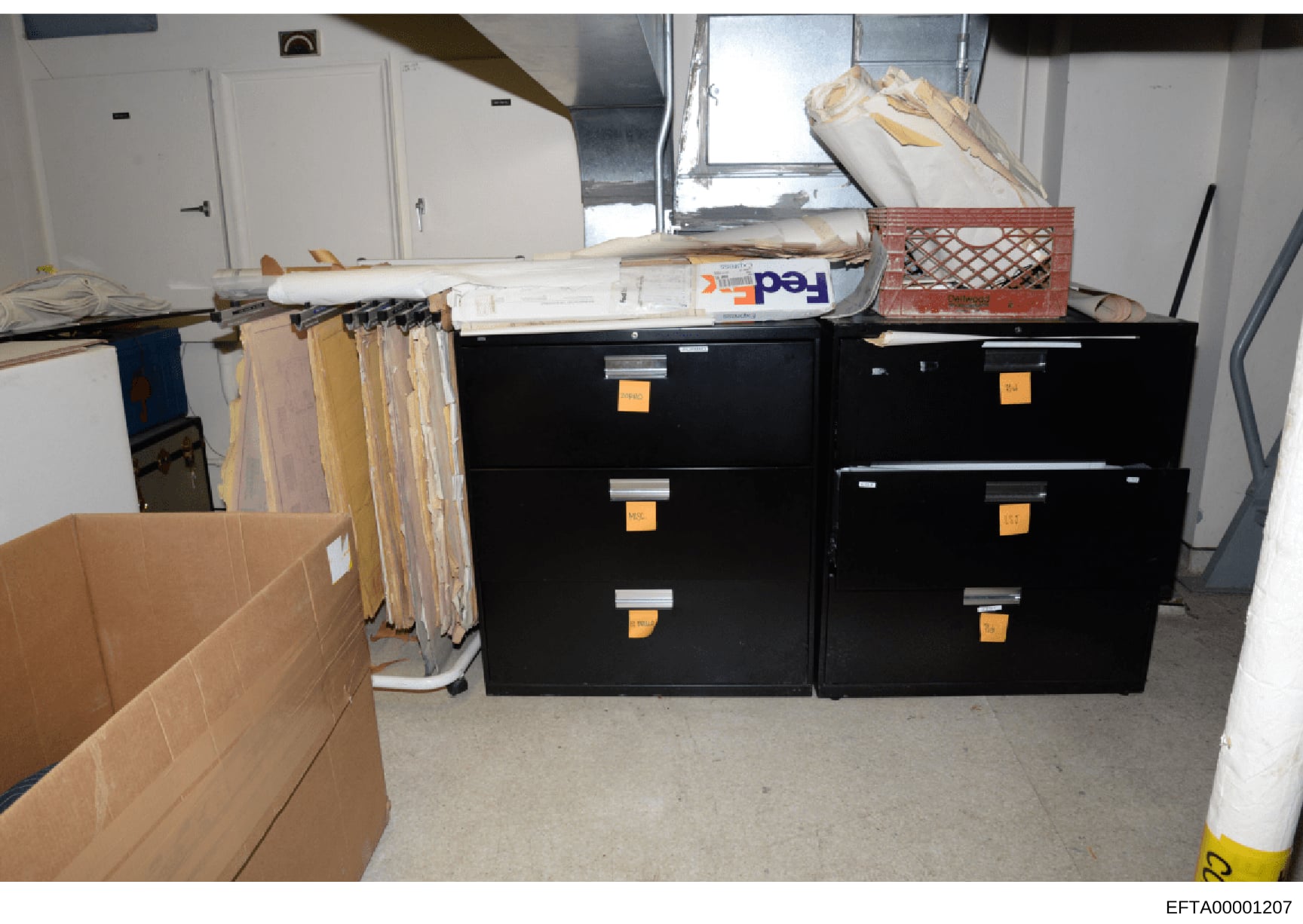 This photograph depicts a storage or utility room containing two large black filing cabinets with orange identification labels, along with various boxes, rolled documents, and other stored materials. The image appears to be evidence documentation fro