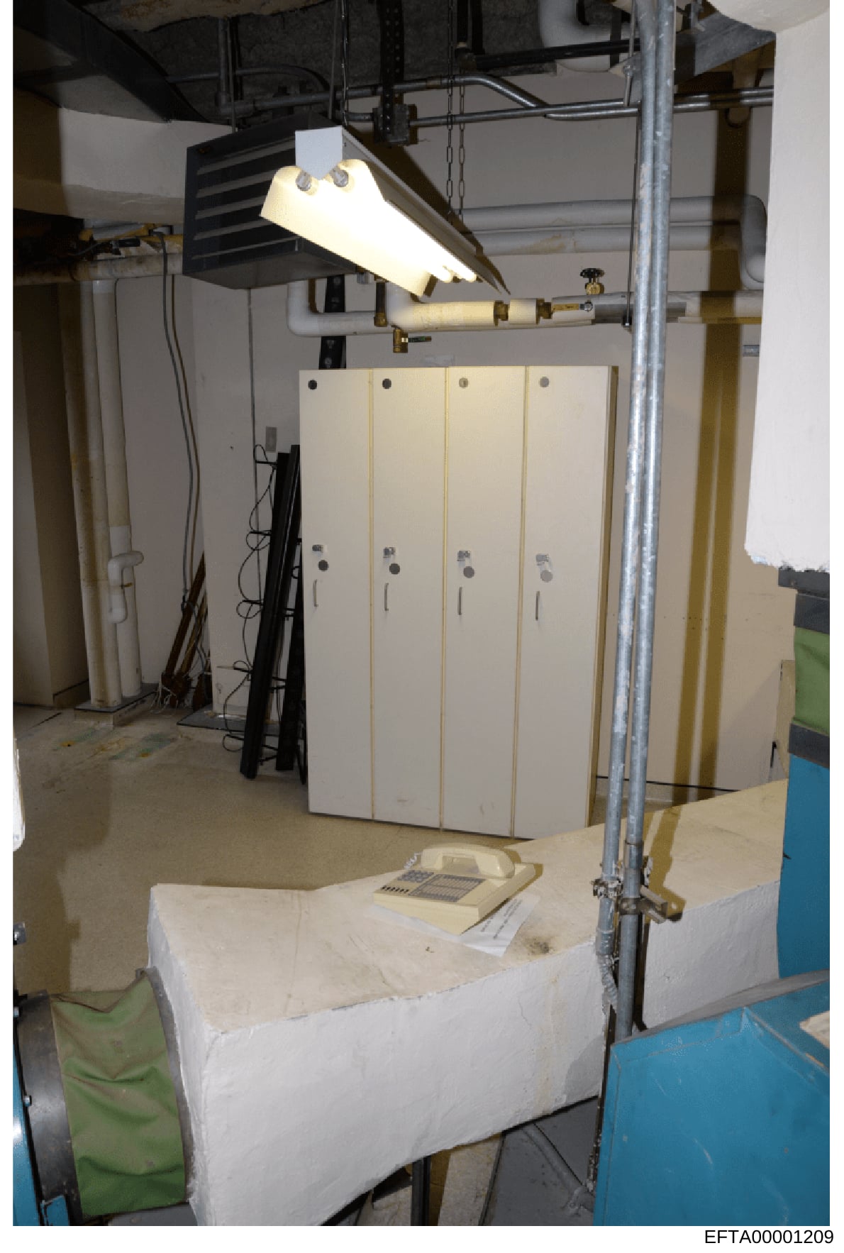 This photograph shows an interior space, likely a basement or utility room, containing four vertical storage lockers or cabinets with locks, industrial lighting fixtures, metal support structures, and various cables and wiring. The room appears to be