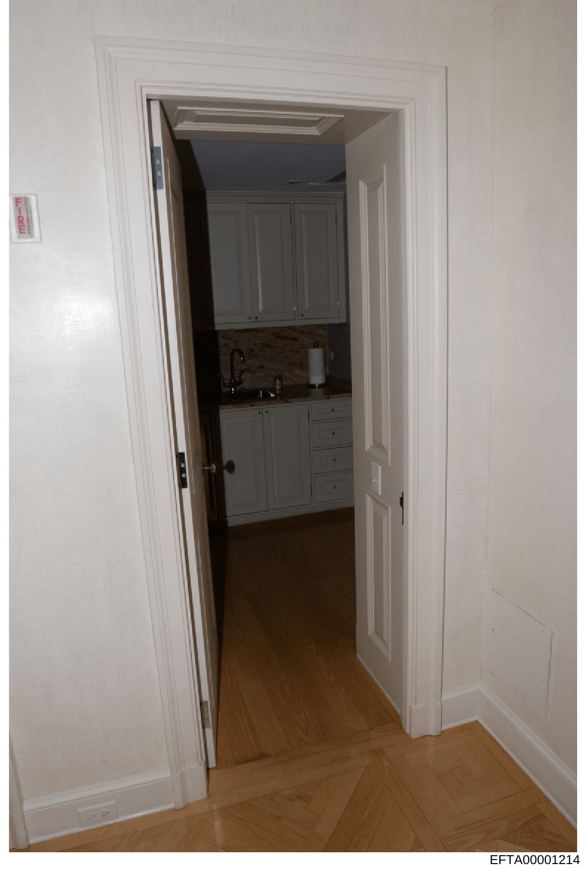 This is a documentary photograph showing the interior of a residential space, specifically a view through a doorway into a kitchen area. The image captures white trim work, wooden cabinetry, countertops, and hardwood flooring. This type of photograph