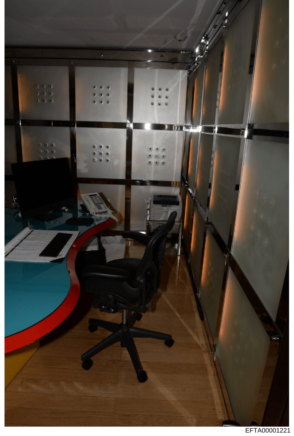 This photograph documents an interior space containing office equipment and storage infrastructure, likely taken as part of the evidence collection process during the investigation. The room features a desk workspace with a black office chair, organi