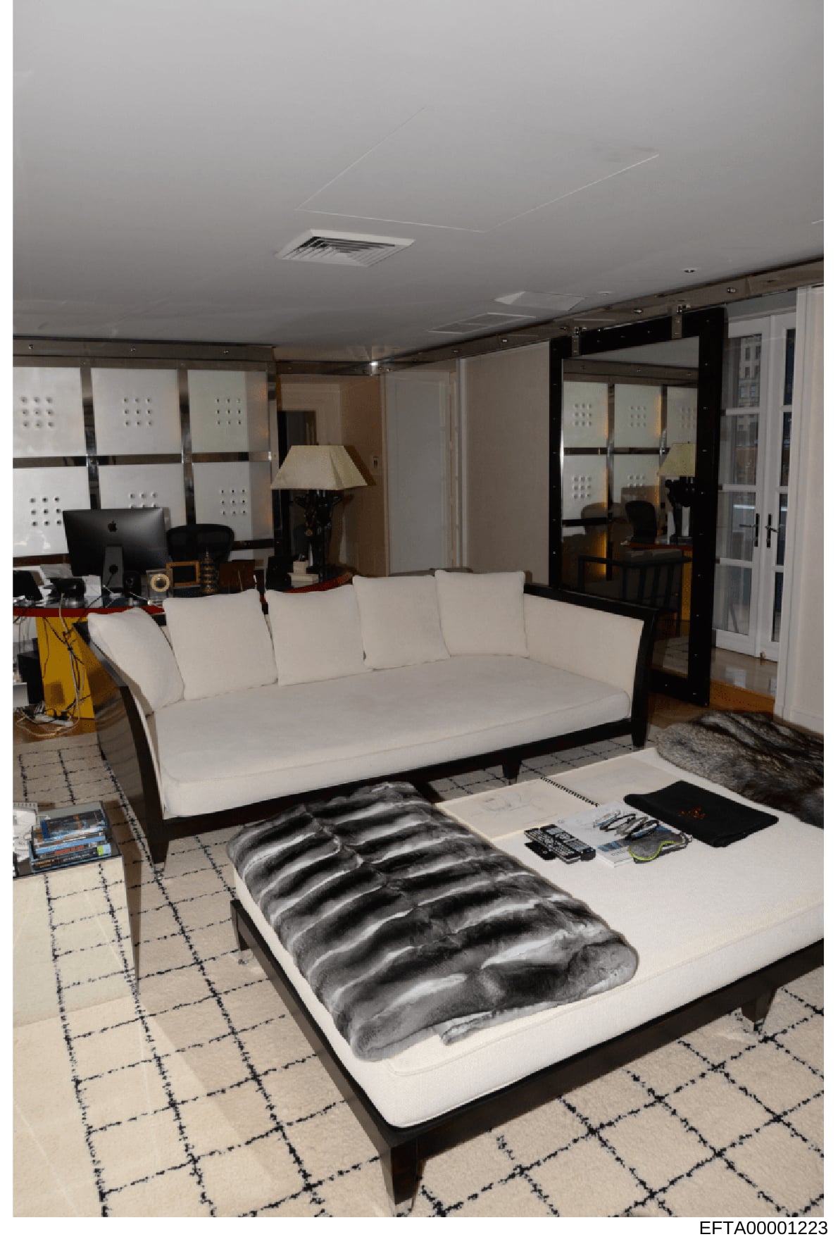This photograph shows a residential living room or lounge area featuring contemporary furniture and décor. The space includes a white upholstered sofa, a bed or daybed with a dark patterned mattress cover, a desk workstation with computer equipment,