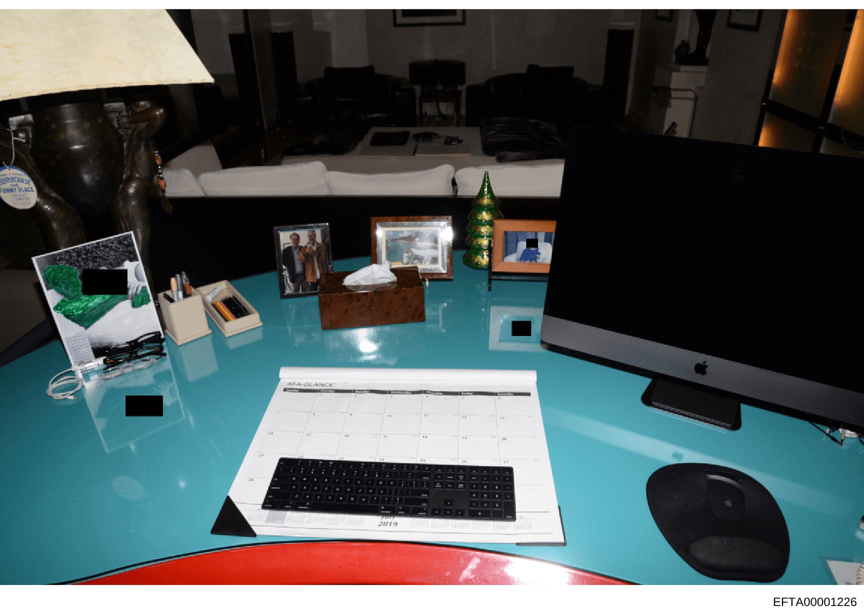 This photograph shows a turquoise desk setup in what appears to be a residential office or study space. The desk contains a computer monitor, keyboard, calendar (dated 2019), framed photographs, decorative items, and various office supplies. The imag