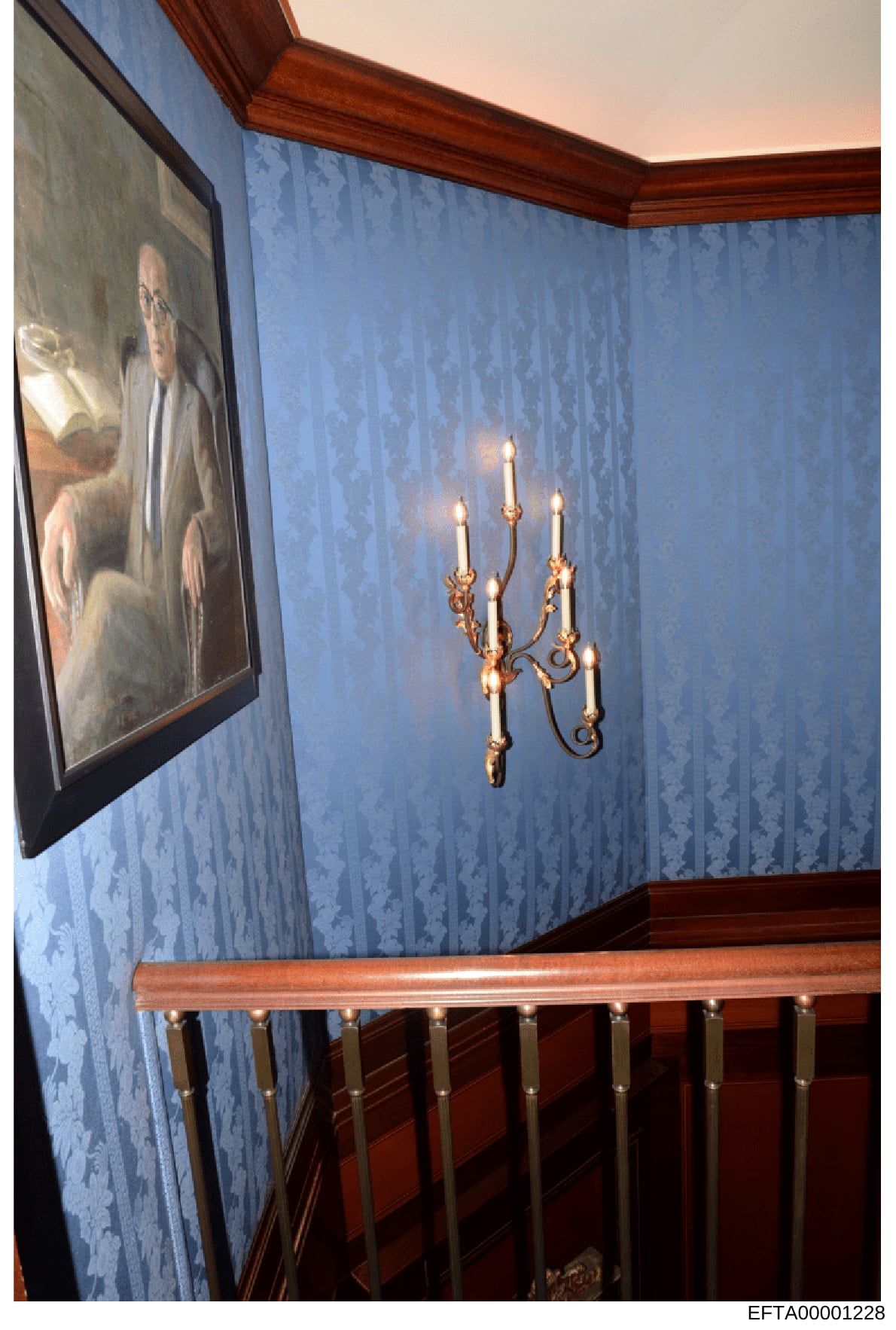 This is a photograph of an interior room space, likely from one of Epstein's properties, showing a corner with blue damask wallpaper, a gold ornate candelabra mounted on the wall, and a framed portrait painting on the left side. The room features dar