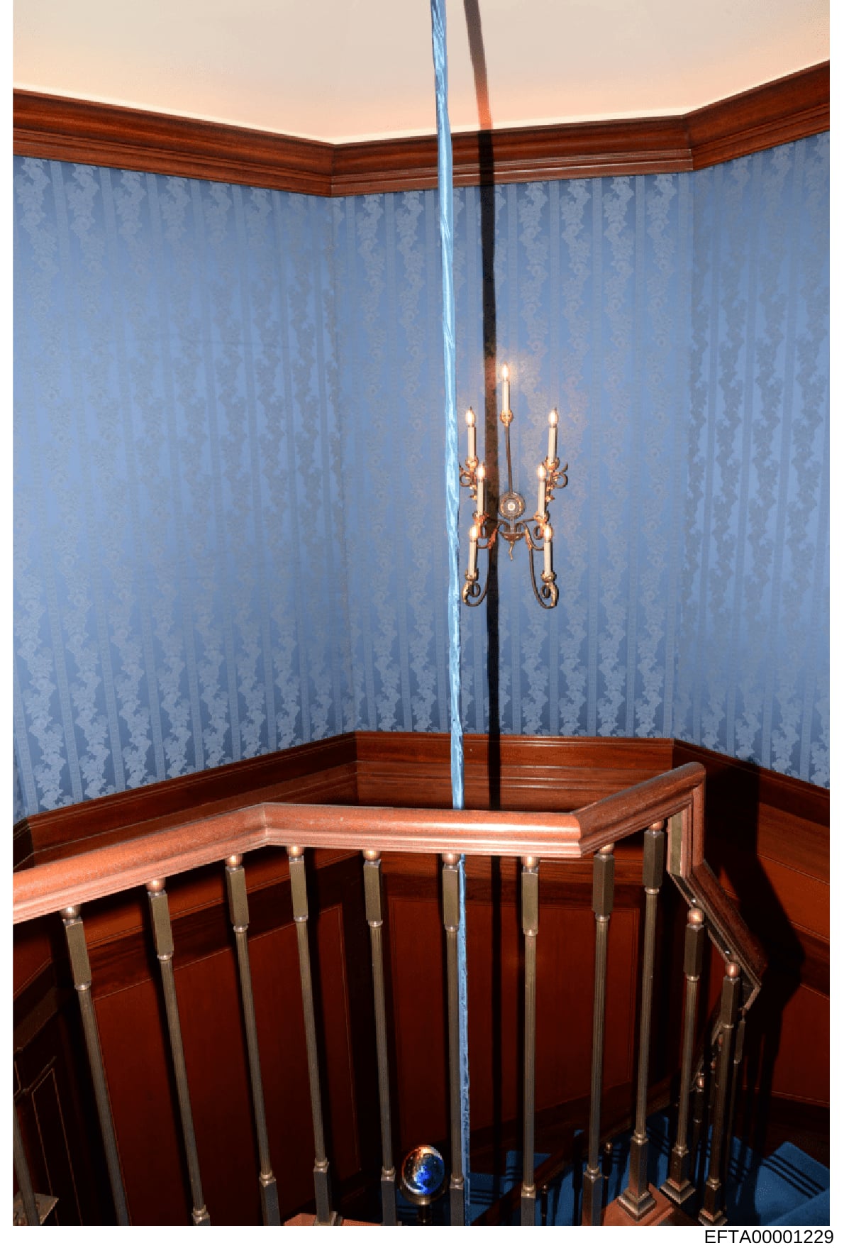 This is an evidence photograph showing the interior of a bedroom featuring a wooden crib with metal railings, decorative blue patterned wallpaper with a damask design, a dark wood wainscoting, and an ornate brass candelabra suspended from the ceiling
