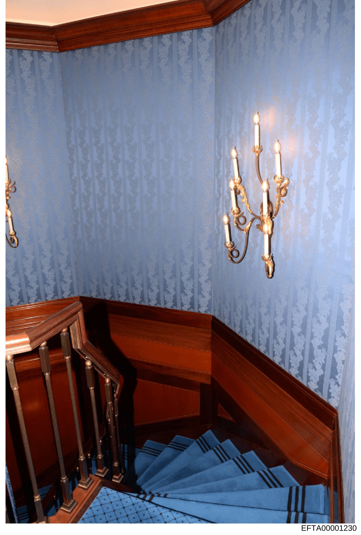 This is a photograph of an interior stairwell featuring elegant classical design elements. The space shows a curved staircase with blue-patterned carpet treads, dark wood railings and wainscoting, light blue damask-patterned wallpaper, a decorative w