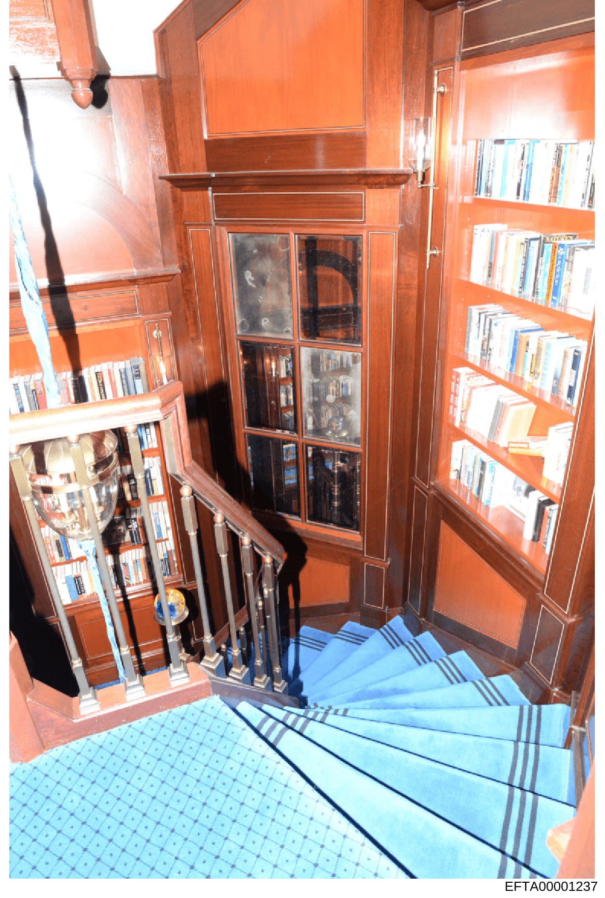 This photograph documents an interior room featuring custom wooden cabinetry with glass-fronted shelving units storing books and media, blue carpeted stairs descending into the space, and warm orange-toned wood paneling. The image appears to be taken
