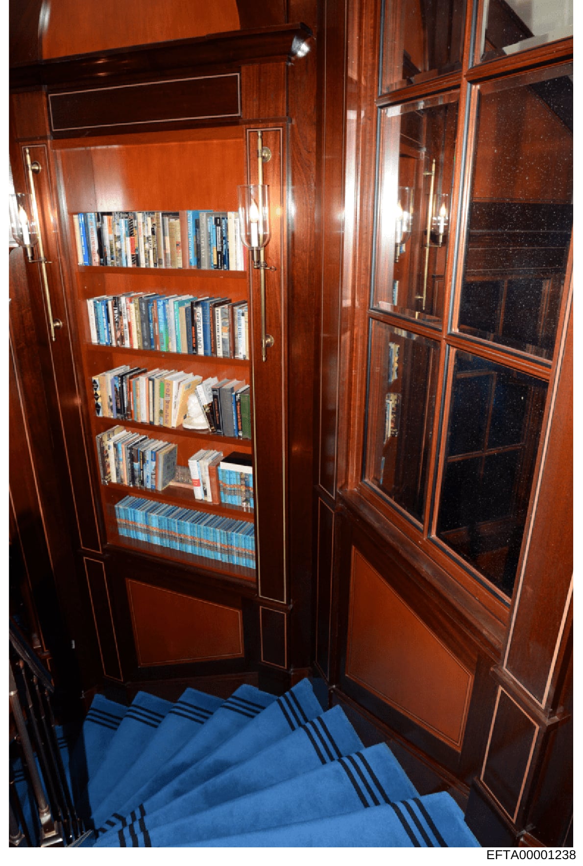 This photograph shows a built-in wooden bookcase with glass-fronted doors containing DVDs, Blu-rays, and books. The image appears to be an inventory or property documentation photo, likely taken during the investigation of an Epstein property. The bo
