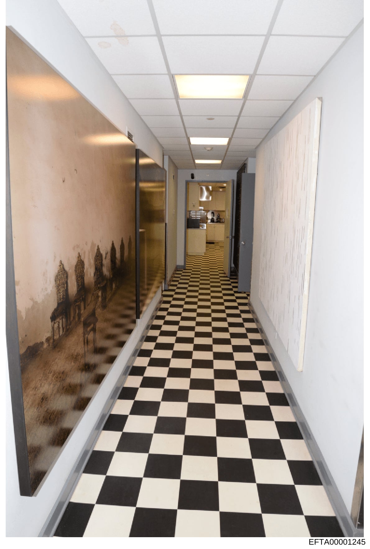This photograph shows a long, narrow hallway with a black and white checkered tile floor, fluorescent ceiling panels, and white walls. On the left wall is artwork depicting what appears to be a cityscape or architectural silhouette. The hallway exten