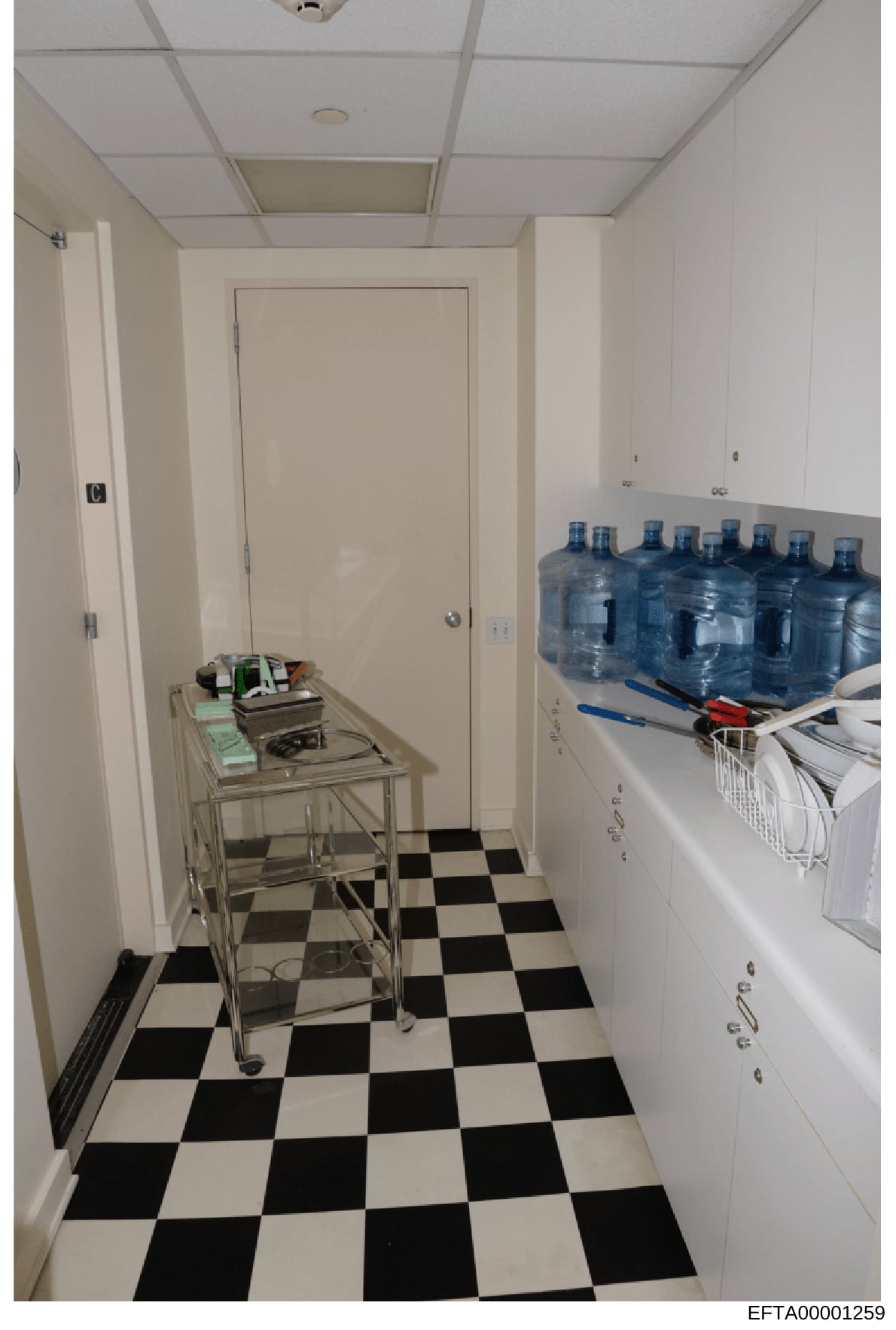 This is a photograph of a residential utility room or pantry space, featuring a black-and-white checkered floor, white cabinetry, a metal utility cart with various items, and multiple large water bottles stored on a countertop. The image appears to b