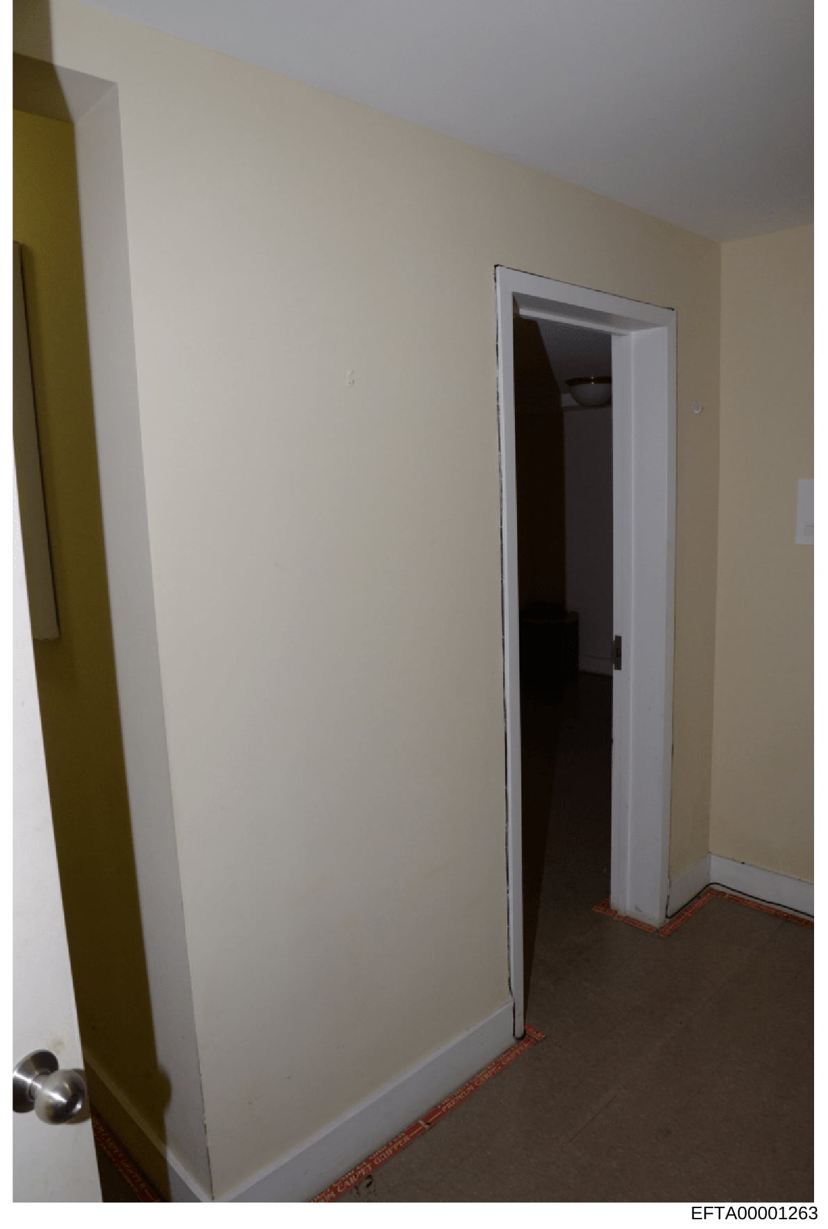 This is a photograph of an interior hallway or corridor space showing cream-colored walls, a white-framed doorway, and dark carpeting with red/orange trim along the baseboard. The image appears to be evidence documentation from a property search or i