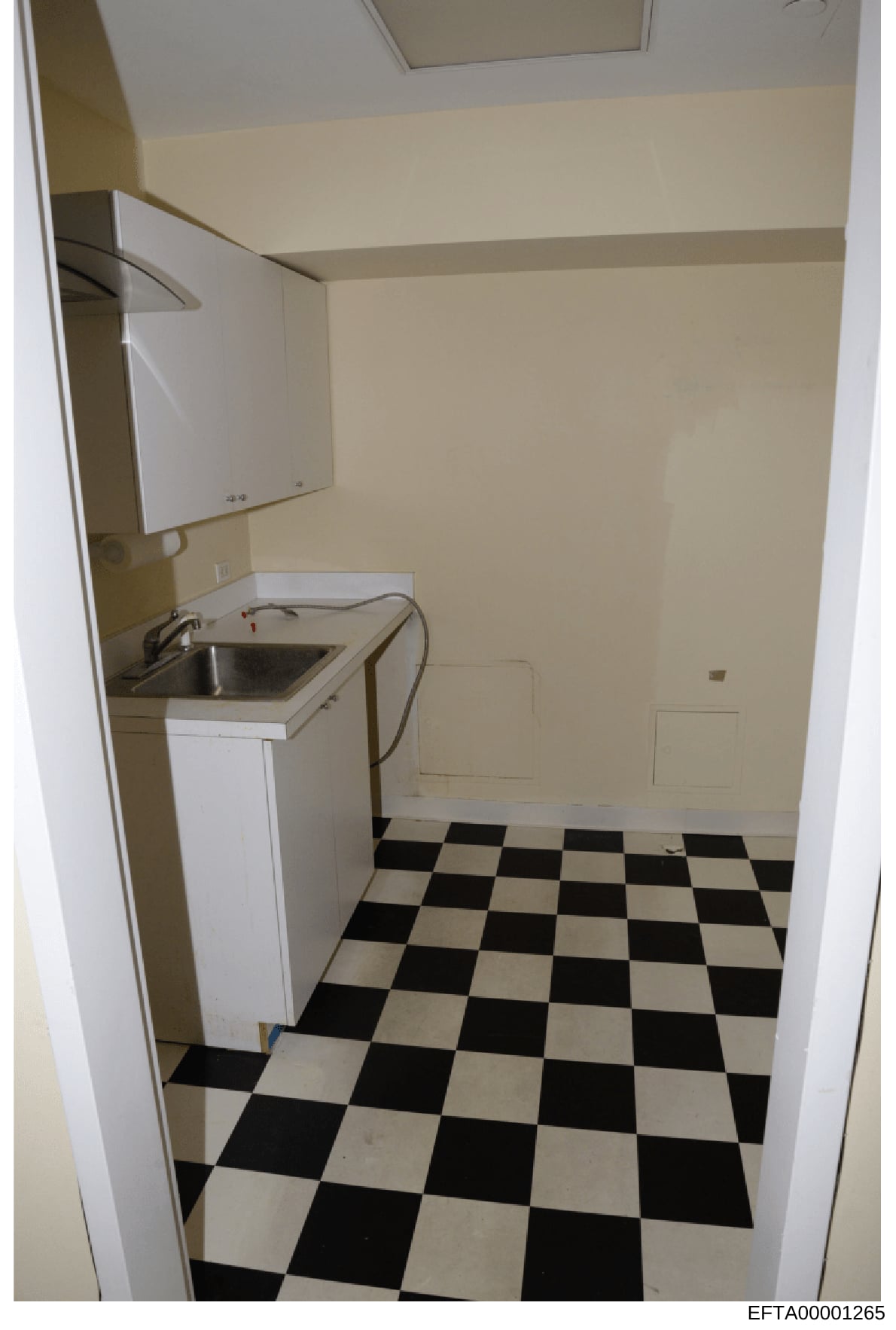 This is a photograph of a small kitchen or utility room interior, showing basic appliances and fixtures. The image appears to be evidence documentation from a property investigation, featuring a compact sink area with overhead cabinet, checkered blac