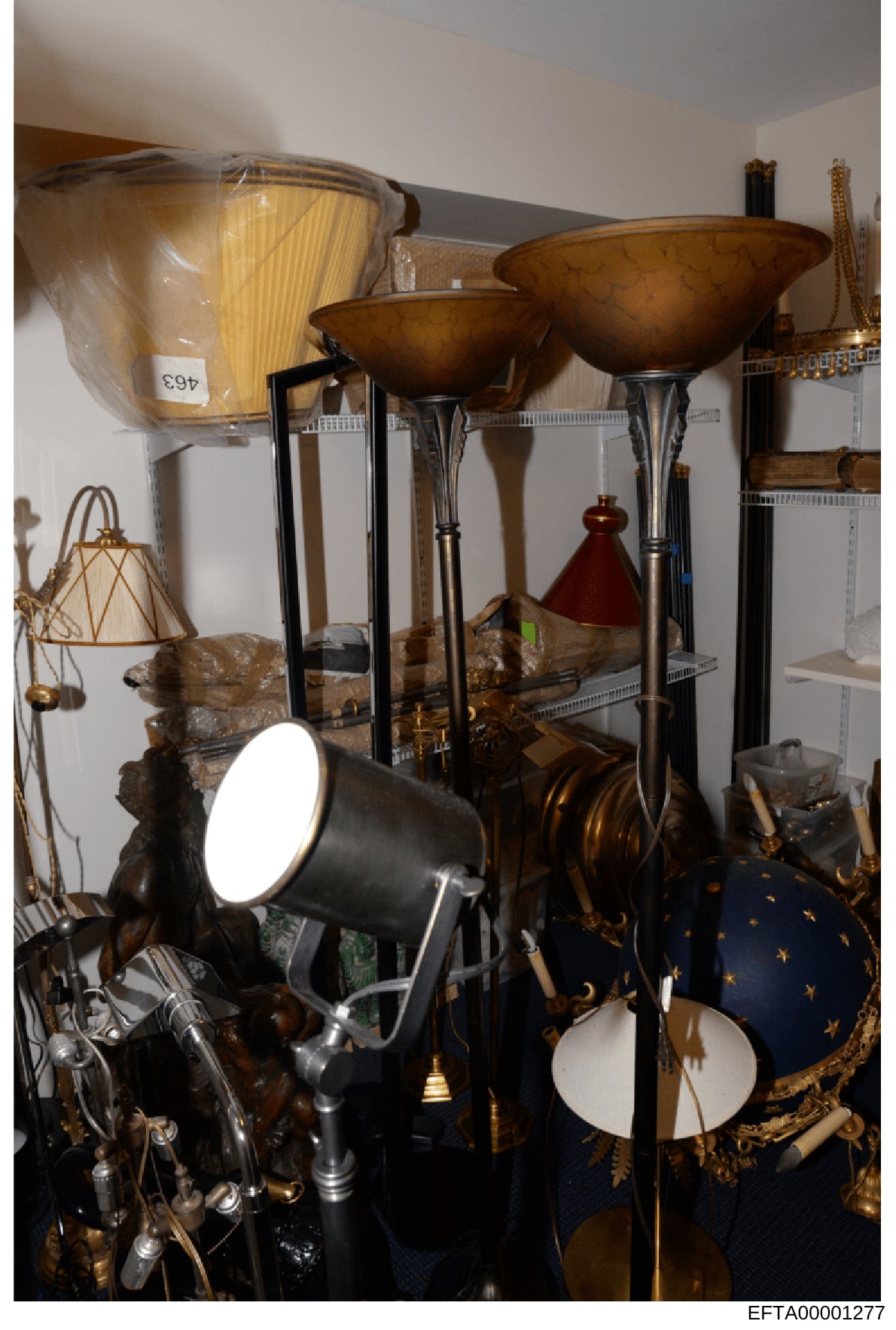 This photograph documents the contents of a storage or display room containing various lighting fixtures, decorative home furnishings, and accessories. The image shows floor lamps with bronze bowl shades, spotlights, woven baskets, ornamental spheres