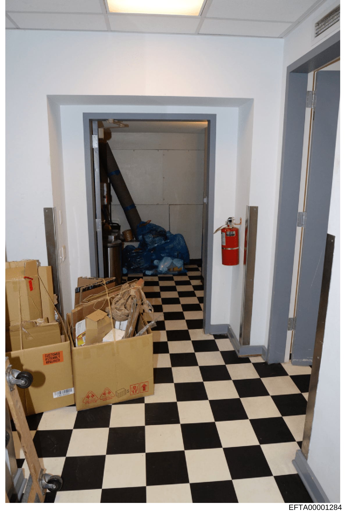 This is an evidence photograph showing an interior storage or utility room with a distinctive black and white checkered floor. The room contains multiple cardboard boxes, blue material/bags, and a red fire extinguisher mounted on the wall. The photog
