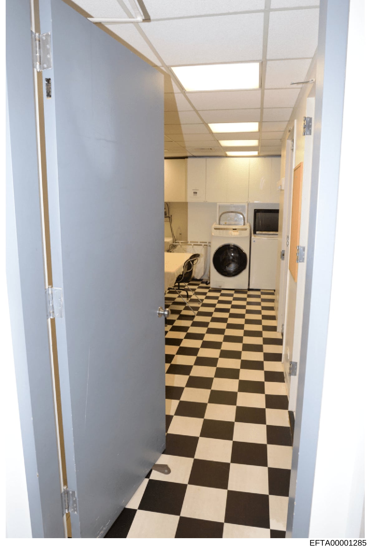 This is a photograph of a utility or laundry room showing domestic appliances including a washing machine, dryer, and sink. The room features a black and white checkered floor and drop ceiling with fluorescent lighting. The image appears to be docume