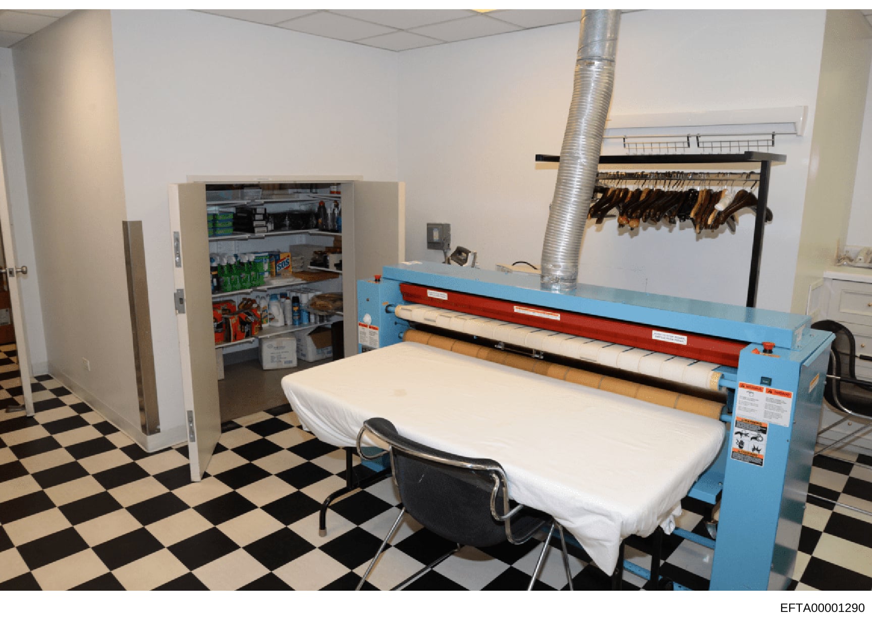 This is a photograph of a utility/laundry room, likely from one of Jeffrey Epstein's properties. The image shows an industrial-scale laundry pressing machine, commercial refrigerator, storage racks, and a black-and-white checkered tile floor. The equ