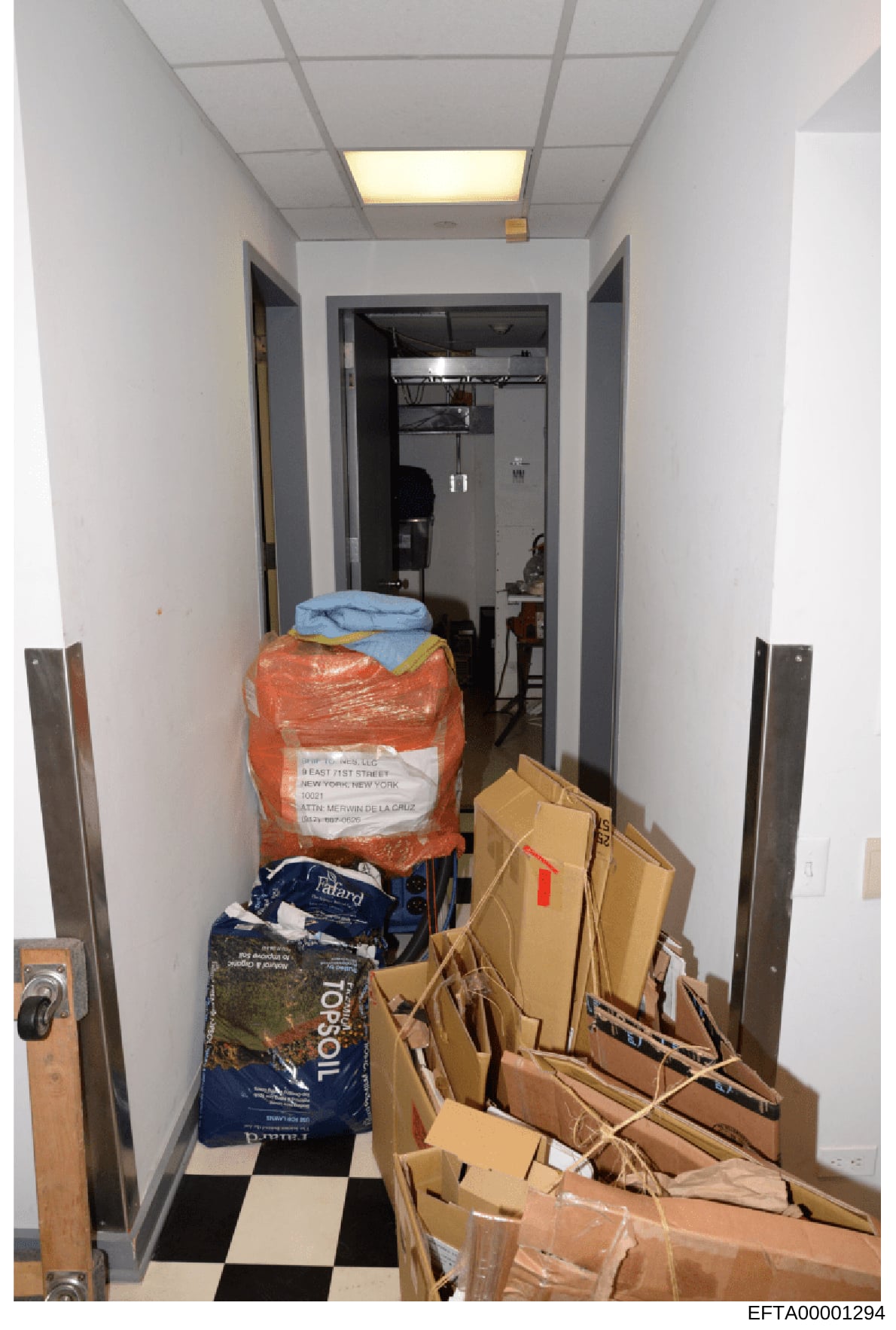 This is an evidence photograph showing the interior of a narrow storage room or closet space containing stacked cardboard boxes, bundled materials wrapped in plastic sheeting, and various household items. The space features white walls, a drop ceilin