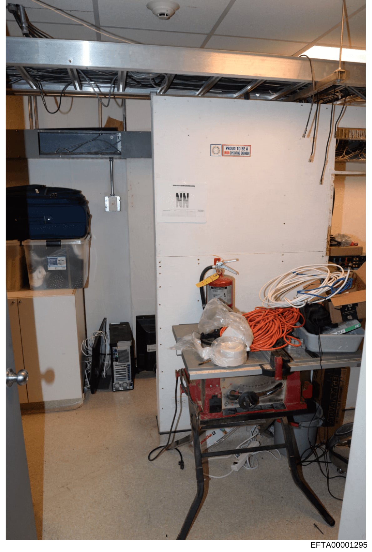 This is an investigative photograph showing the interior of a basement storage room containing various electrical equipment, tools, and materials. The room features exposed ceiling infrastructure, electrical panels, fire safety equipment, coiled cabl