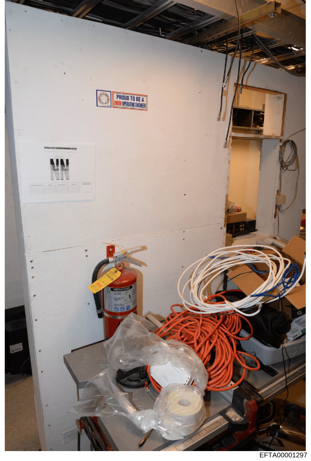 This photograph shows an interior room or workspace, likely in a construction or industrial setting, containing safety equipment including a fire extinguisher, electrical cords, and various tools. A 'Proud to be a Union Operating Engineer' sign is vi
