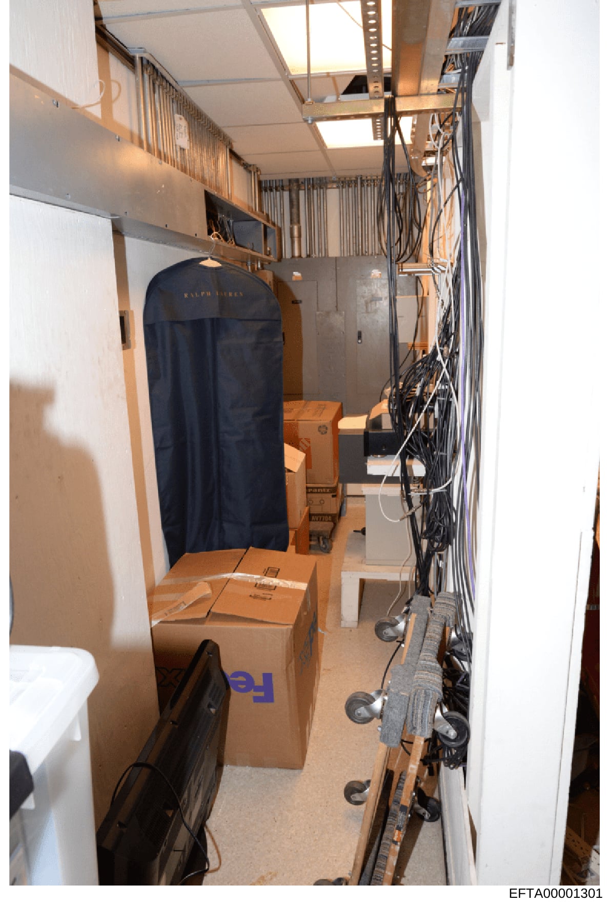 This photograph shows a narrow storage or utility room containing cardboard boxes (including one marked with 'FedEx'), a dark Ralph Lauren garment bag, various cables and electrical equipment, and storage shelving. The room appears to be part of a re