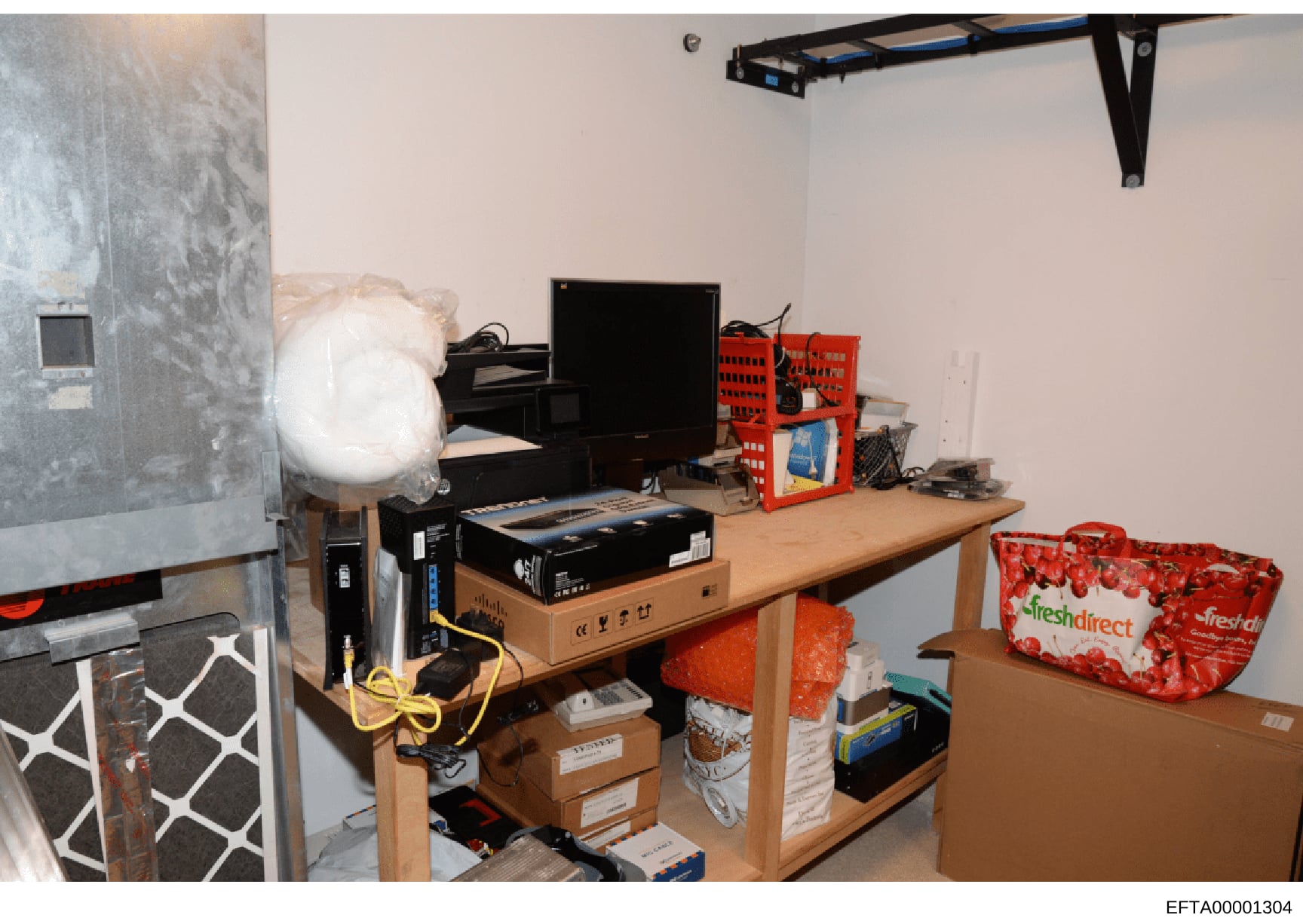 This is an evidence photograph showing an interior room containing computer equipment, networking hardware, storage boxes, and household items. The image appears to be from a search or seizure operation, documented with a Bates number for investigati