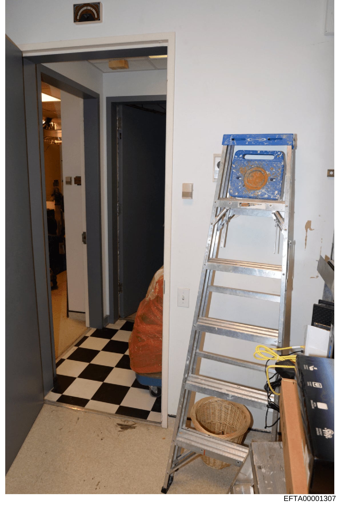 A photograph showing an interior room or hallway with a black and white checkered floor, an open doorway leading to another space, and a metal ladder positioned against the right wall. Various household items and electrical equipment are visible. The