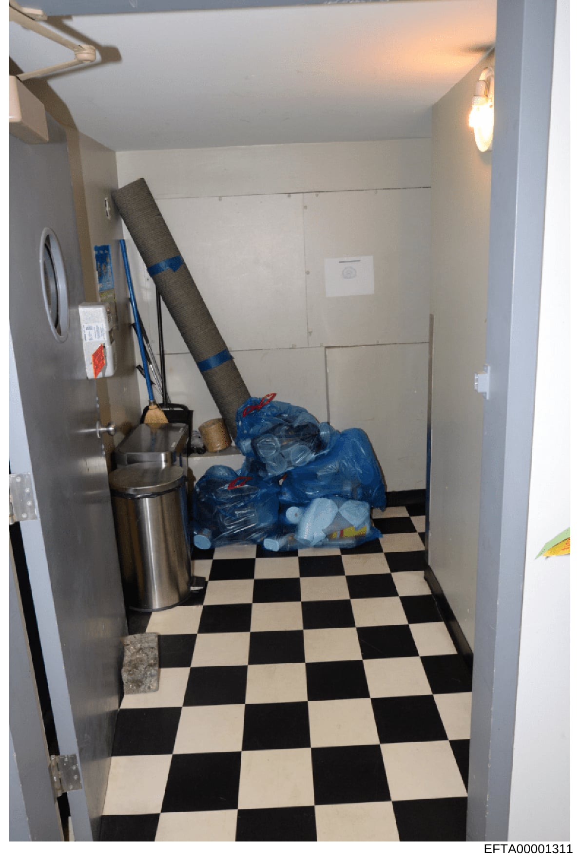 This is an evidence photograph of a kitchen or utility room interior, showing a stainless steel refrigerator, checkered black-and-white flooring, white walls, and stored items including rolled carpeting and blue plastic bags. The photo was taken as p