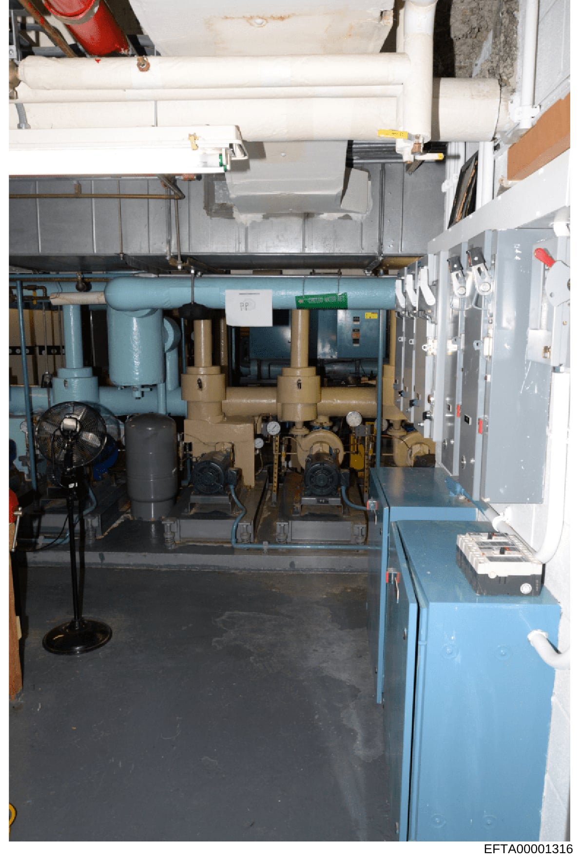 This photograph documents a mechanical/utility room containing HVAC equipment, including large blue and tan-colored compressors or air handling units, piping systems, electrical panels, and various supporting infrastructure. The image appears to be e