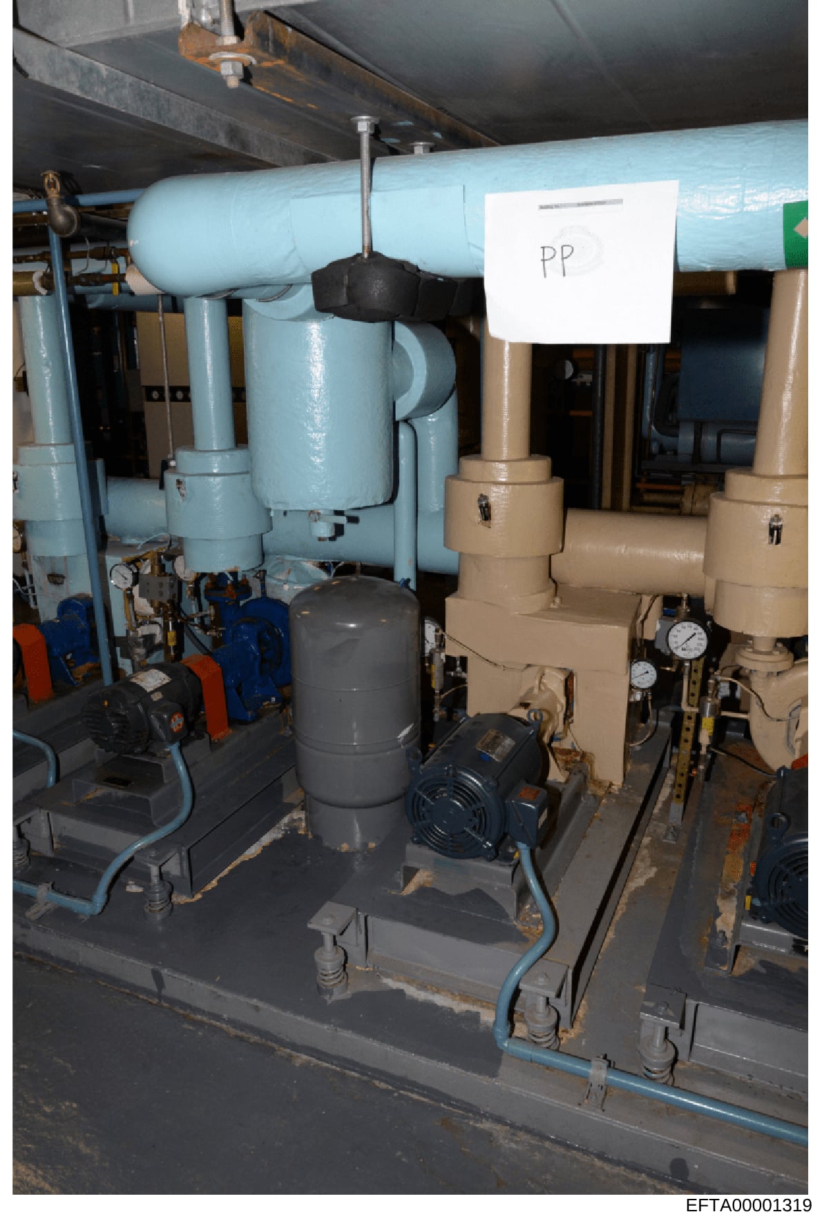 This is a photograph of industrial mechanical equipment, likely part of a building's HVAC or plumbing system. The image shows blue and tan-colored pipes, pumps with motors, gauges, and a black valve or connector. A white label marked 'PP' is visible 