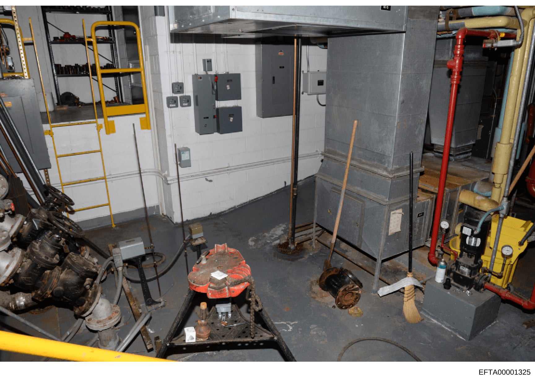 This is a photograph of an industrial or maintenance workspace containing various mechanical equipment, electrical panels, hydraulic systems, and tools. The image shows a basement or ground-level utility room with multiple large machines, electrical 
