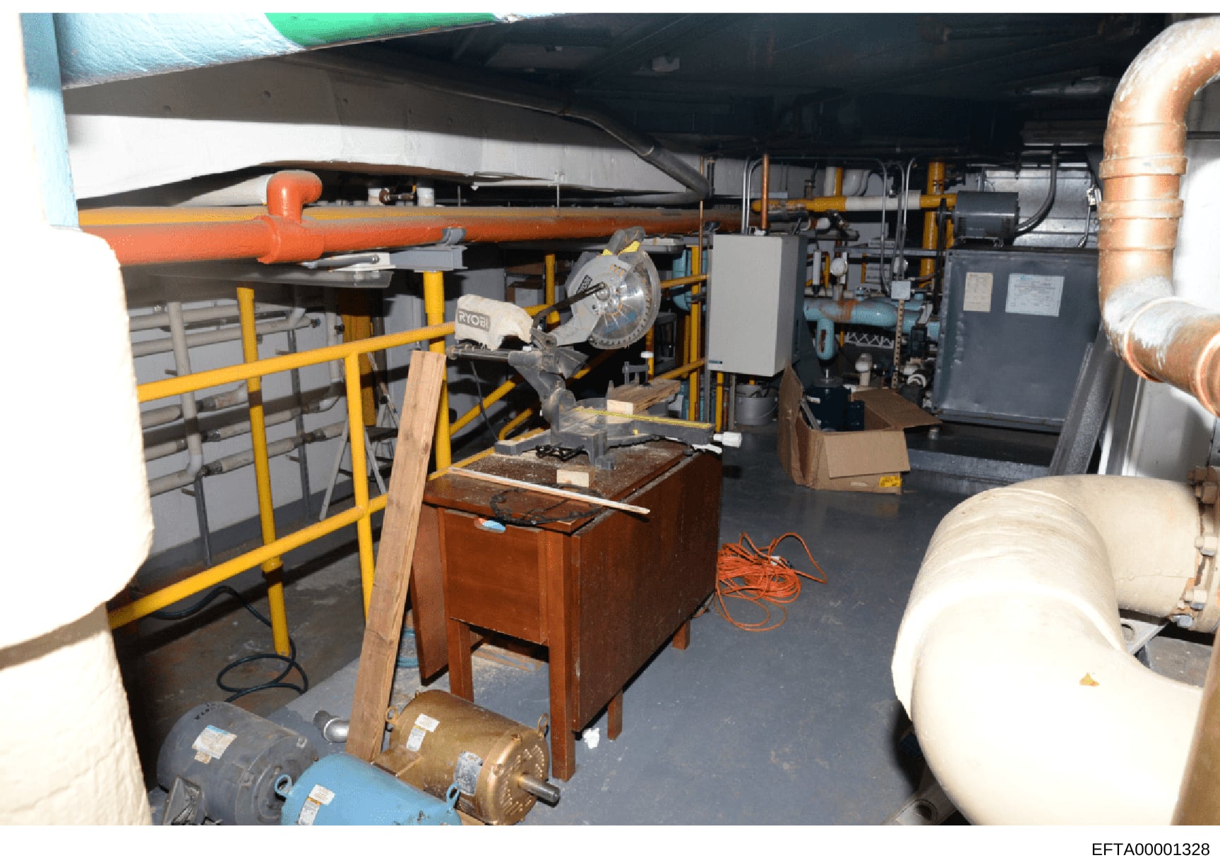 This is a photograph of a mechanical room or basement area containing HVAC equipment, piping systems, and various building infrastructure. The image shows industrial equipment including an orange pipe, yellow safety railings, copper piping, electrica