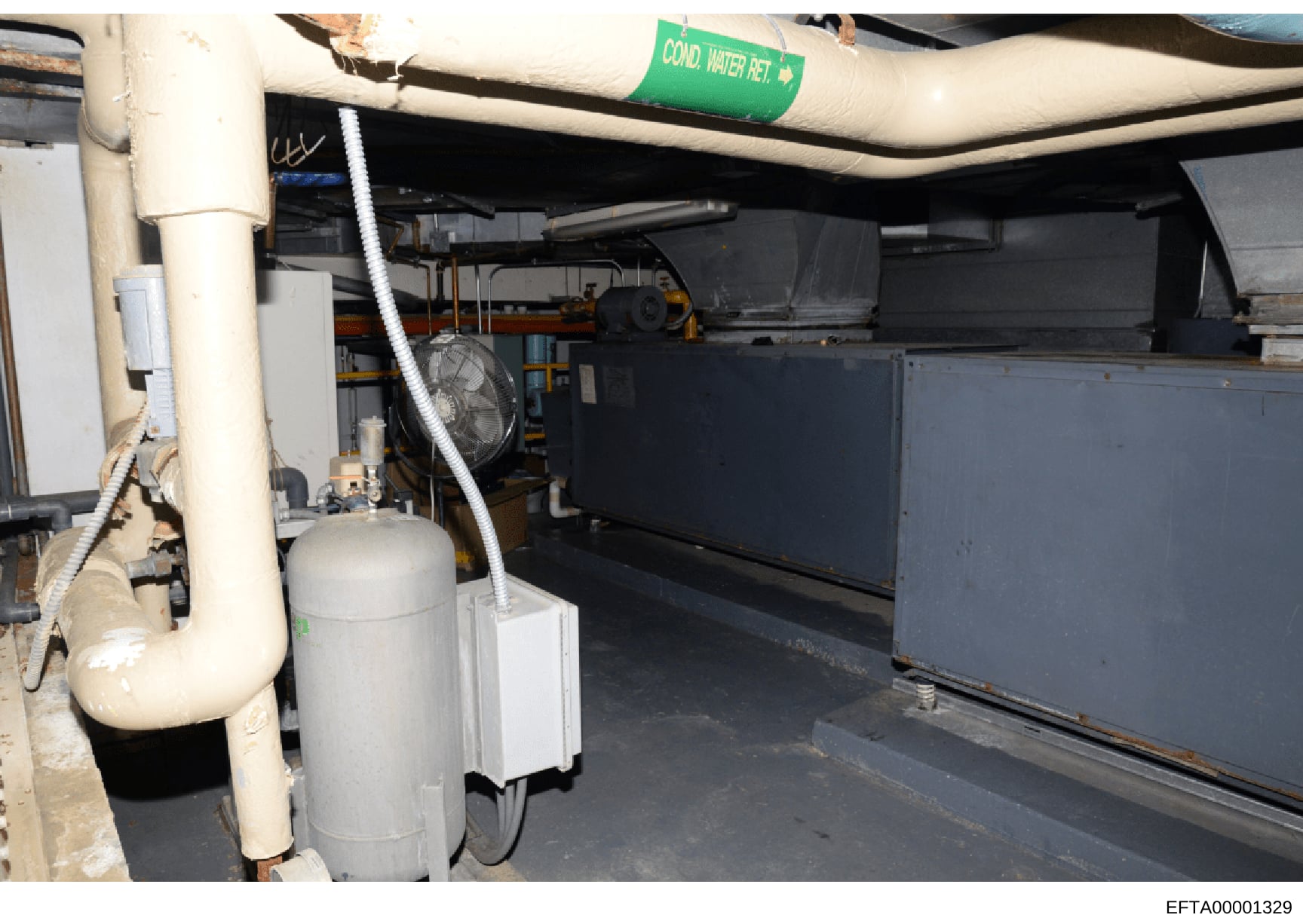 Photograph of a basement or mechanical room showing HVAC systems, plumbing infrastructure, and building utilities. A green label marked 'COND. WATER RET.' (condensation water return) is visible on overhead piping. The image documents the interior mec