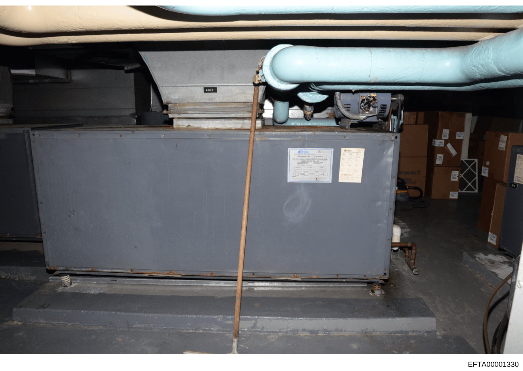 This photograph shows industrial HVAC (heating, ventilation, and air conditioning) equipment installed in what appears to be a basement or mechanical room. The image captures a large gray metal unit with piping, ductwork, and various mechanical compo