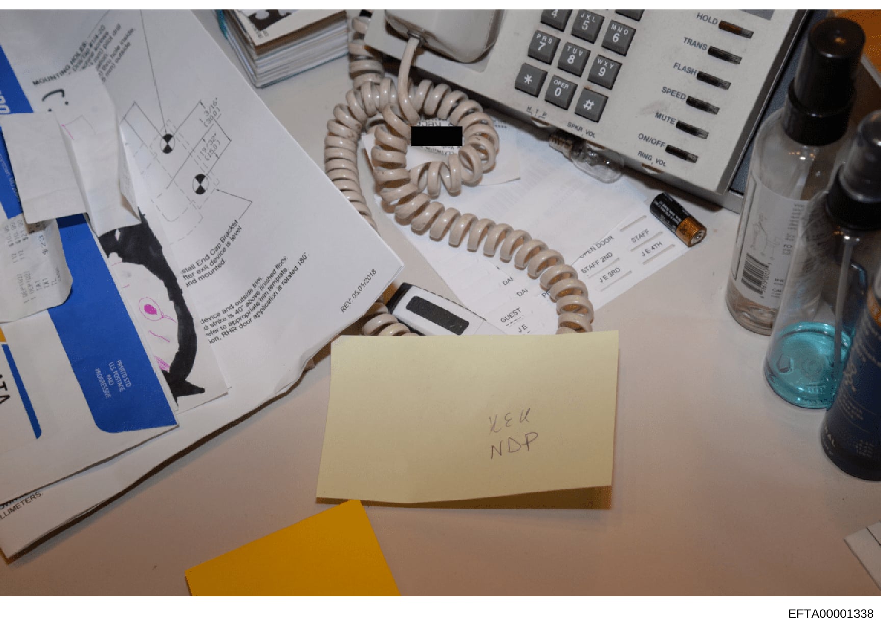 This is a photograph of a workspace or desk surface containing various documents, office equipment, and materials. A yellow sticky note is visible with handwritten text reading 'U&U NDP'. The image shows scattered papers with printed text and diagram