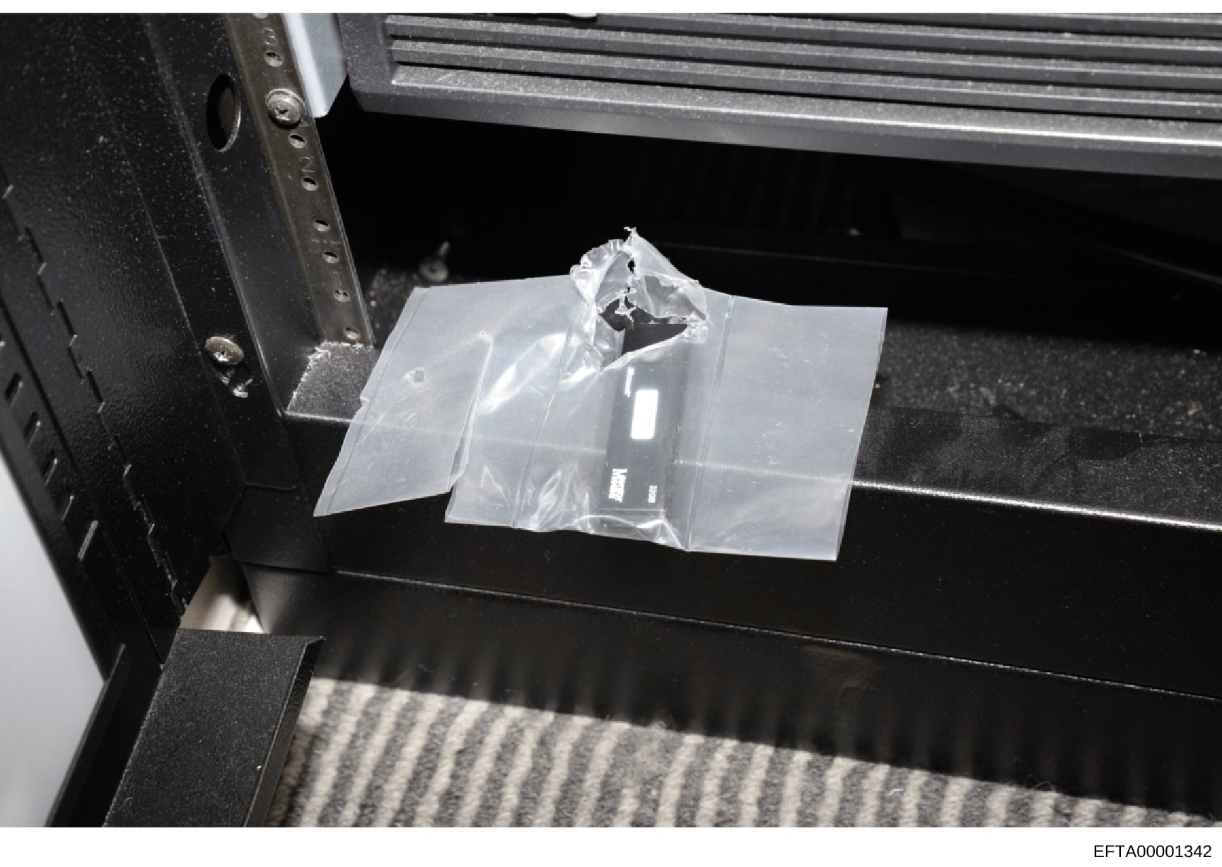 This is a photograph of a small item sealed in a clear plastic evidence bag, positioned on what appears to be a black evidence collection table or surface. The item's specific nature is obscured by the packaging and angle of the photograph. The image