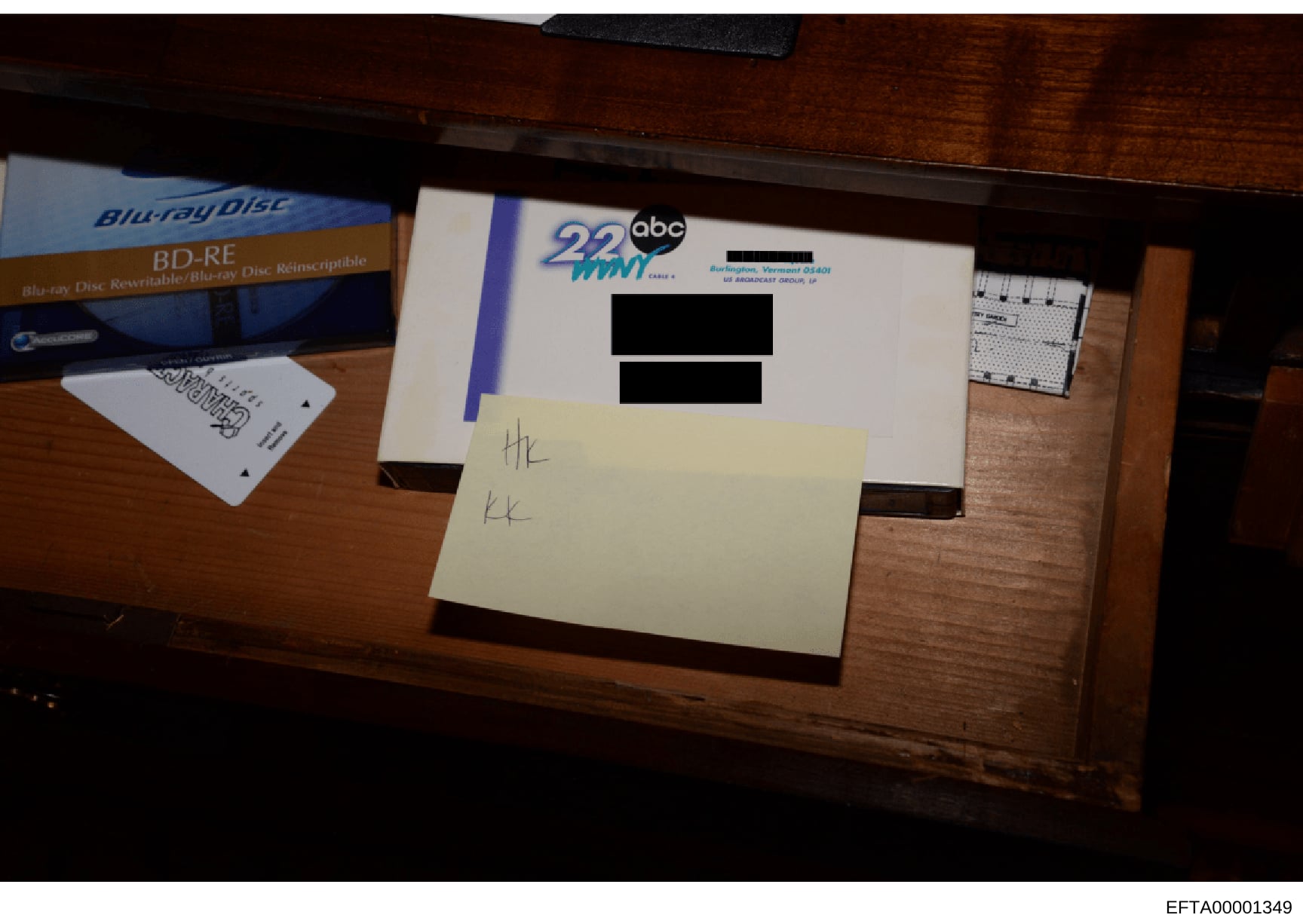 This is an evidence photograph showing a wooden desk surface containing a Blu-ray disc case, what appears to be a television station logo (ABC 22 WNM), and handwritten notations on a yellow sticky note. The image includes redacted text on a white doc