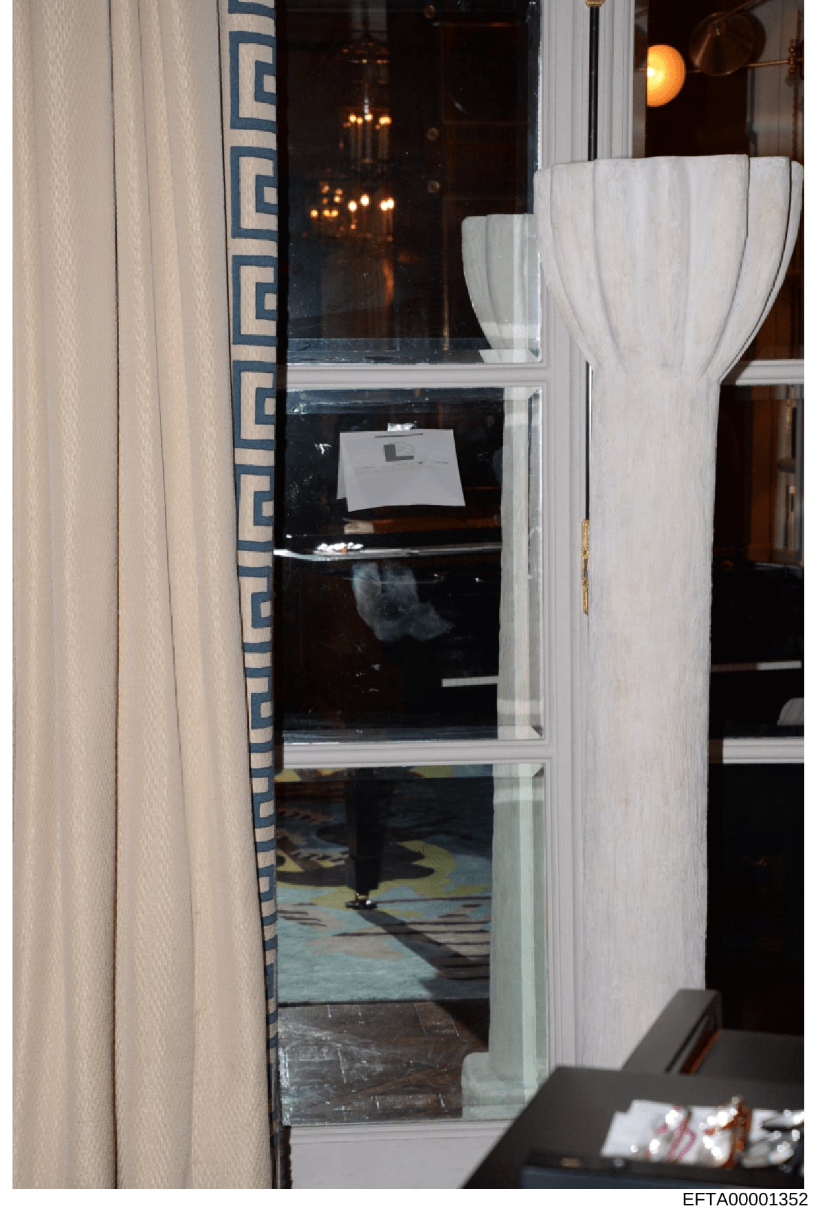 This is an evidence photograph showing the interior of a room taken through a glass door or window. The image captures decorative furnishings including white curtains, patterned upholstery with blue geometric designs, and interior lighting fixtures. 