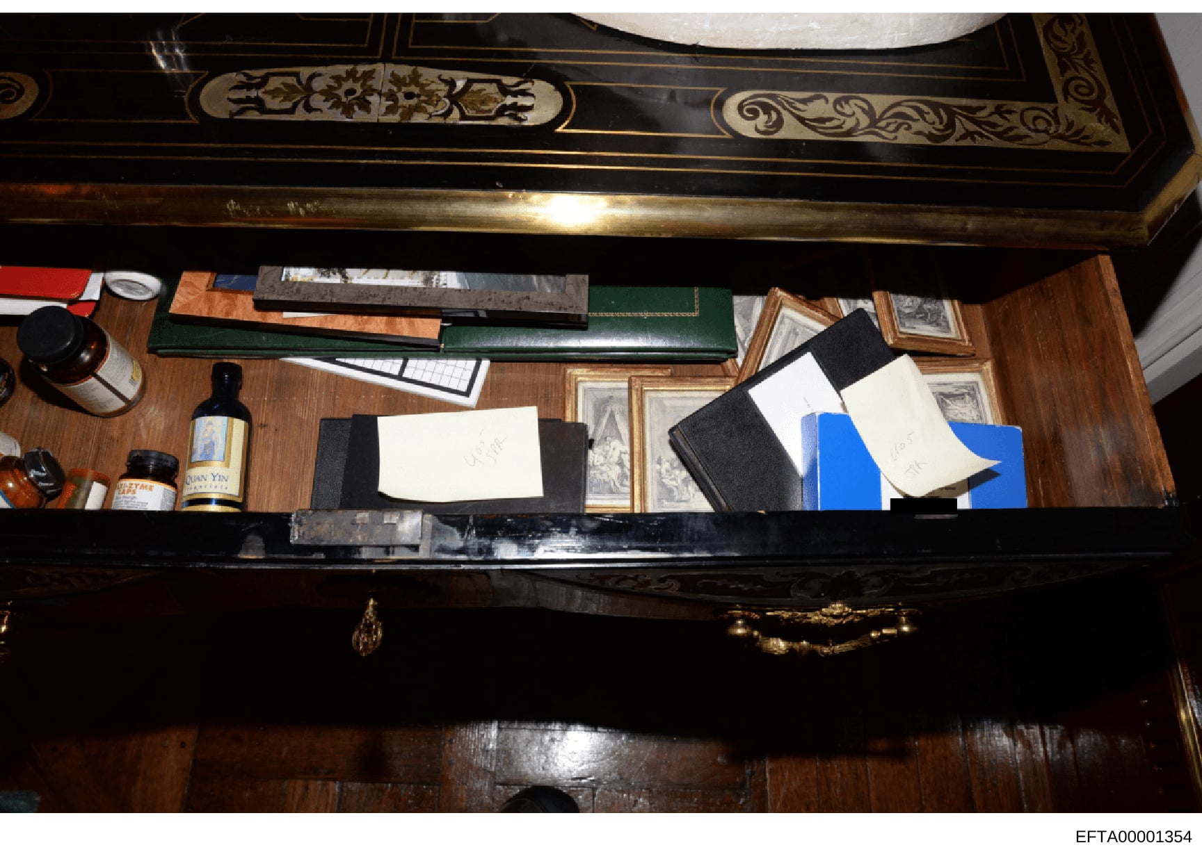 This photograph shows the interior of an ornate desk with decorative inlaid wood and brass details. The desk contains various documents in folders (including green and black leather folders), framed photographs, bottles of spirits, and scattered pape