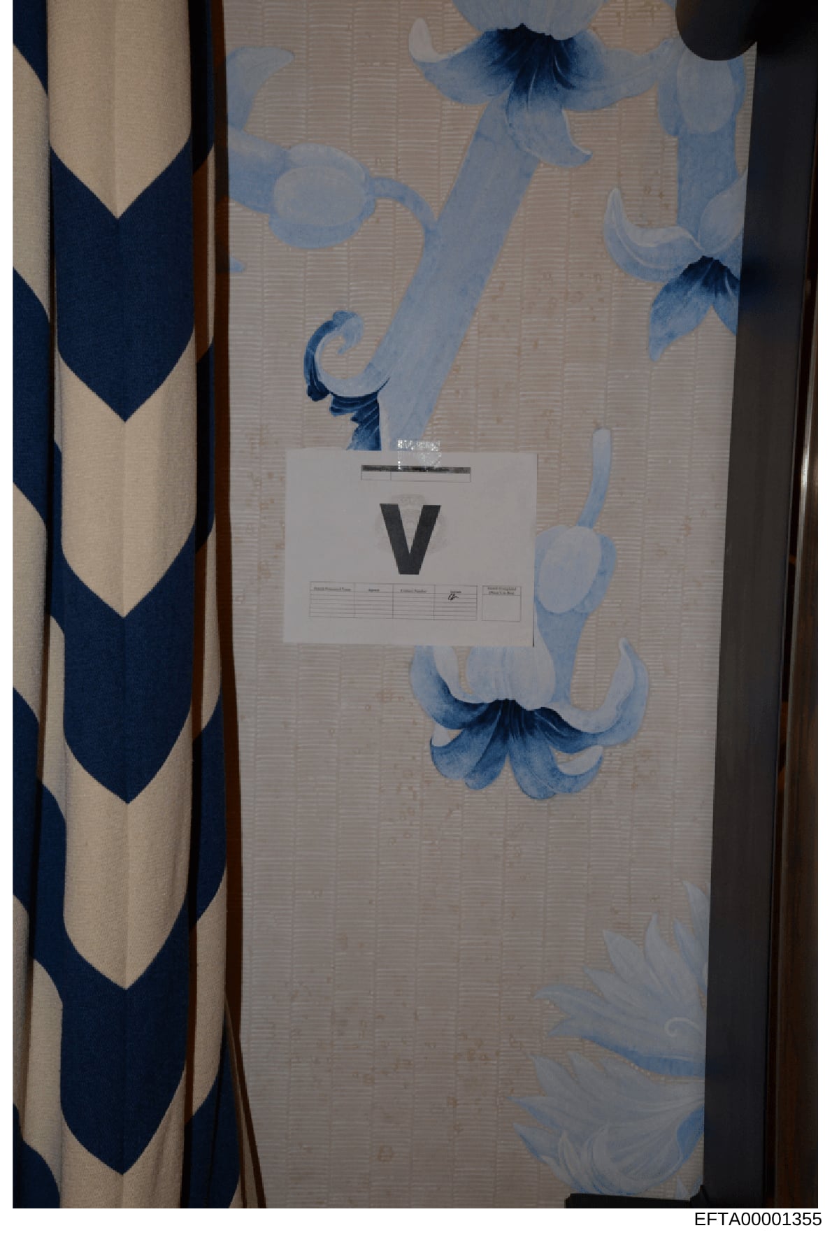 This is an evidence photograph showing an interior wall or surface with decorative blue and white floral wallpaper or fabric pattern. A white evidence marker labeled 'V' is placed on the surface for documentation and scale reference. The image includ