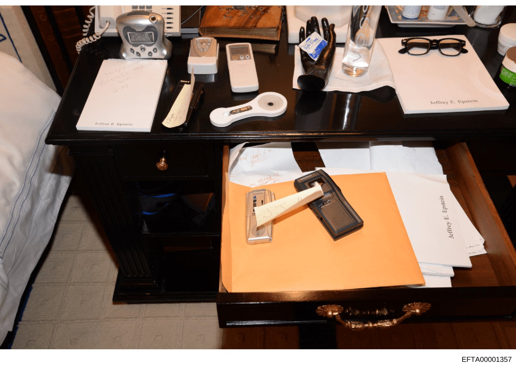 This is an evidence photograph documenting the contents of Jeffrey Epstein's desk, seized during the investigation. The image shows a dark wooden desk with multiple drawers containing personal electronics, documents with Epstein's name, writing imple