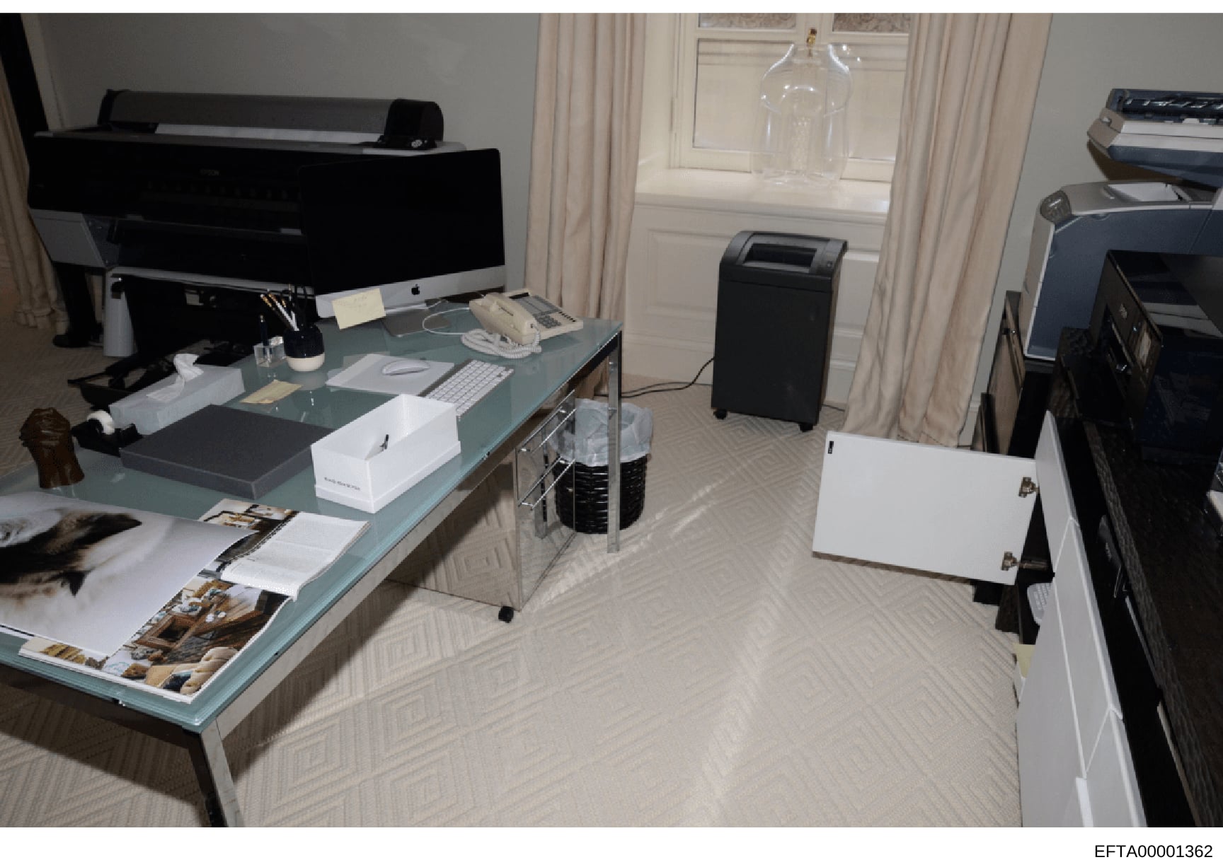 This photograph depicts an office or workspace containing multiple computer workstations, printers, and various personal items and photographs scattered on a glass-topped desk. The image appears to be evidence documentation from a property search or 