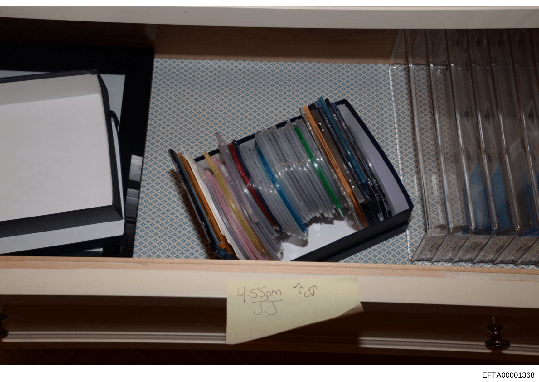 This is a photograph of a shelf containing numerous CDs organized in storage cases with colored spines, along with other media storage materials. A handwritten label reading '4:55pm JJ CD' is visible on the shelf edge, suggesting this photo was taken