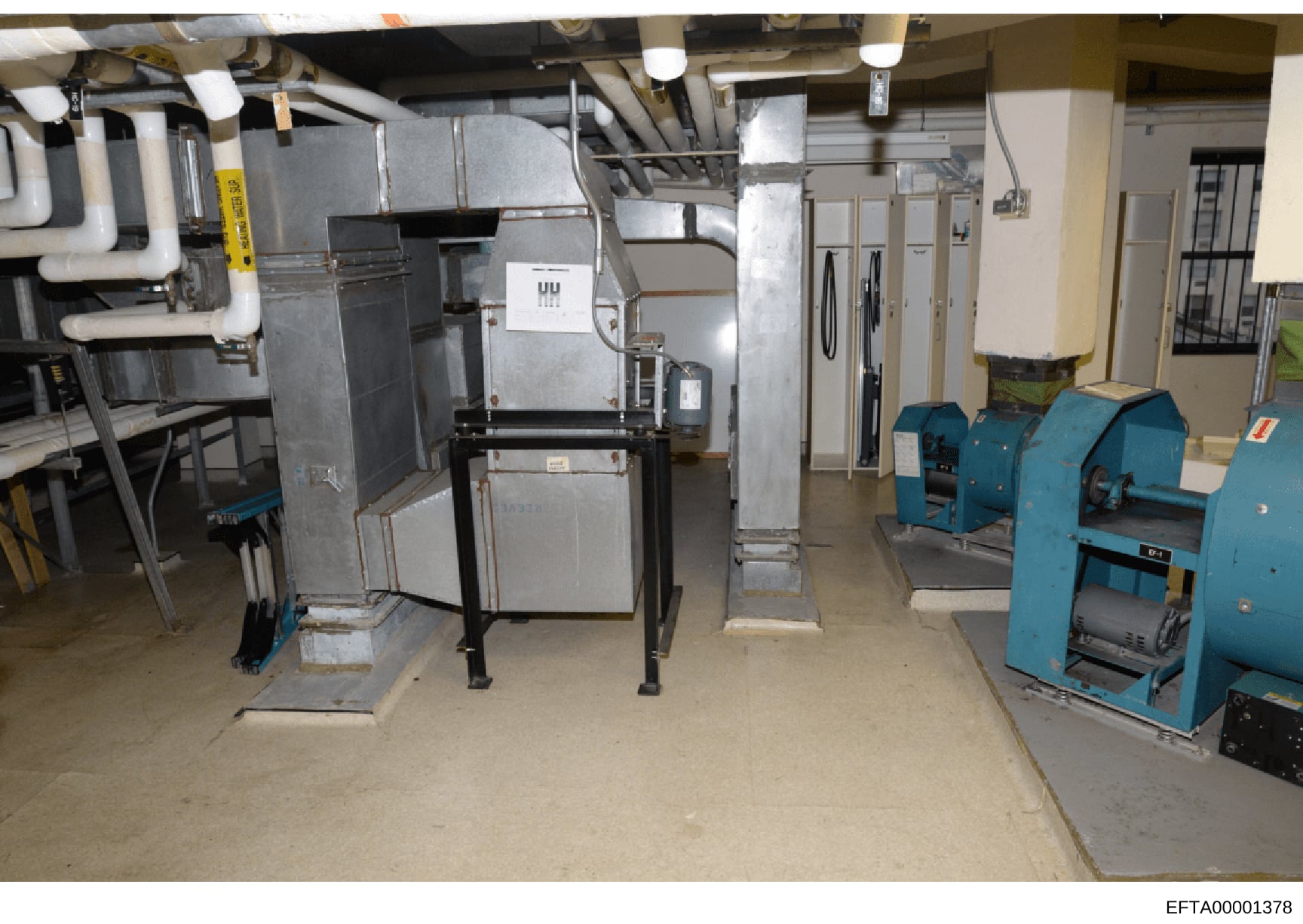 This photograph shows an interior basement or mechanical room containing industrial equipment including HVAC systems, metal ductwork, large metal units (possibly furnaces or air handlers), and blue industrial machinery. The room appears to be part of