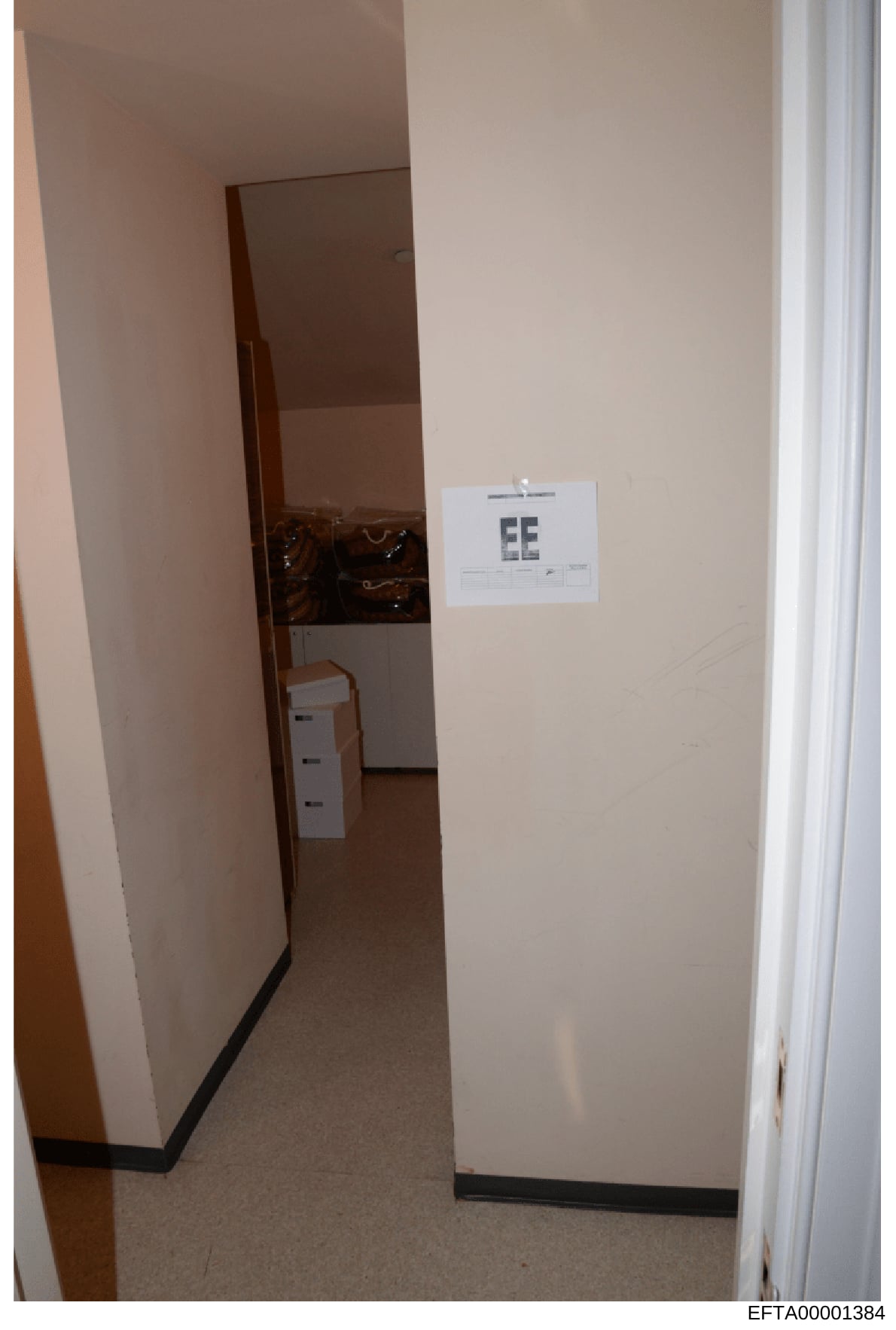 This is a photograph of an interior room or hallway, showing a doorway with white walls, carpeted flooring, and a white label marked 'EE' affixed to the wall. The image appears to be evidence documentation from the investigation, with a Bates number 