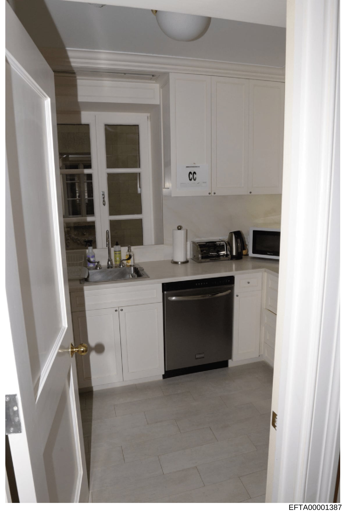 This is a color photograph of a modern, compact kitchen with white cabinetry, stainless steel appliances, and light tile flooring. The image shows a well-maintained residential kitchen space with standard furnishings and countertop items. The photo i