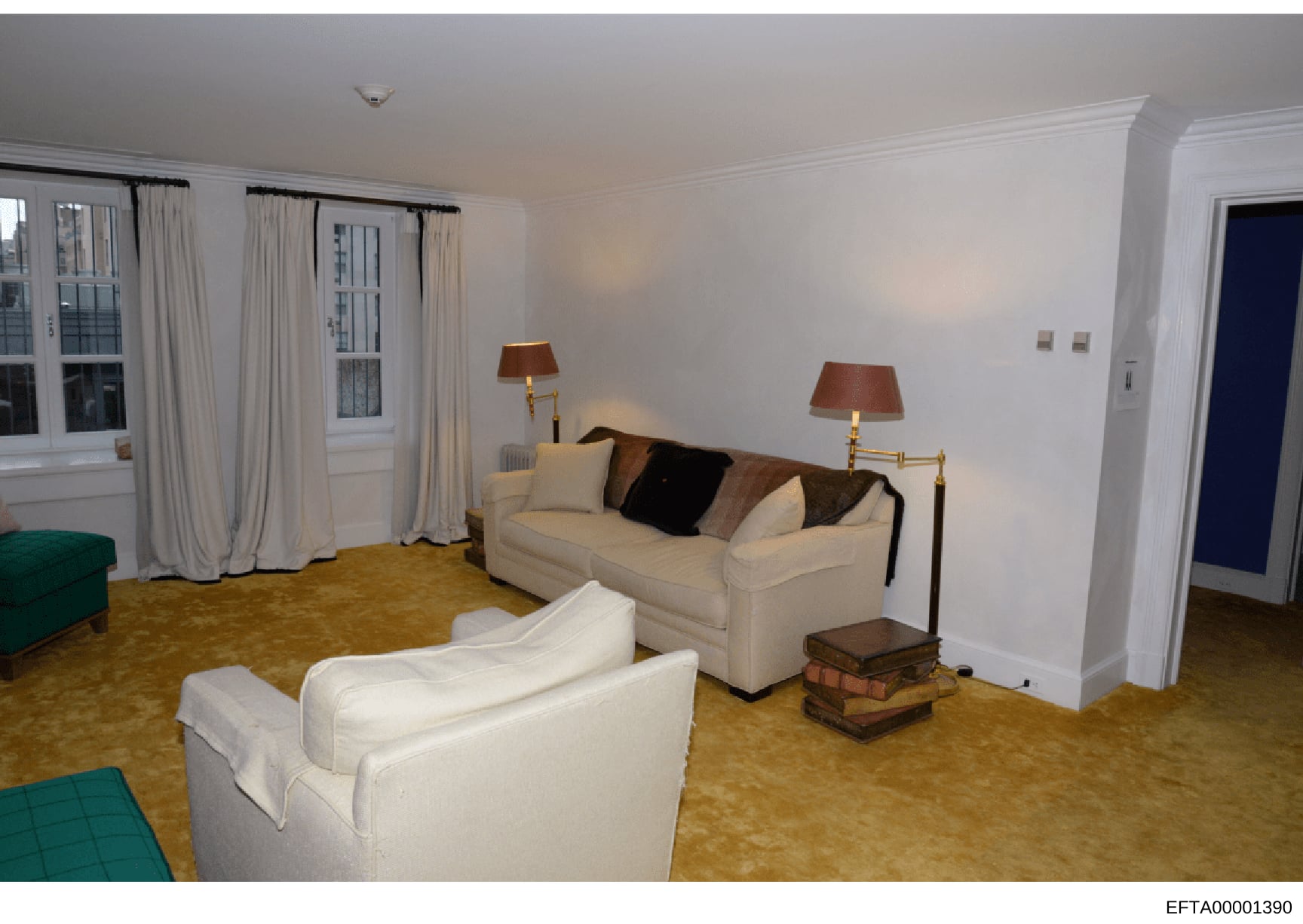 This photograph depicts a luxury residential living room, likely from one of Jeffrey Epstein's properties. The room features high-end furnishings including cream-colored upholstered sofas, brass table lamps, and a gold-toned carpet. Large windows wit