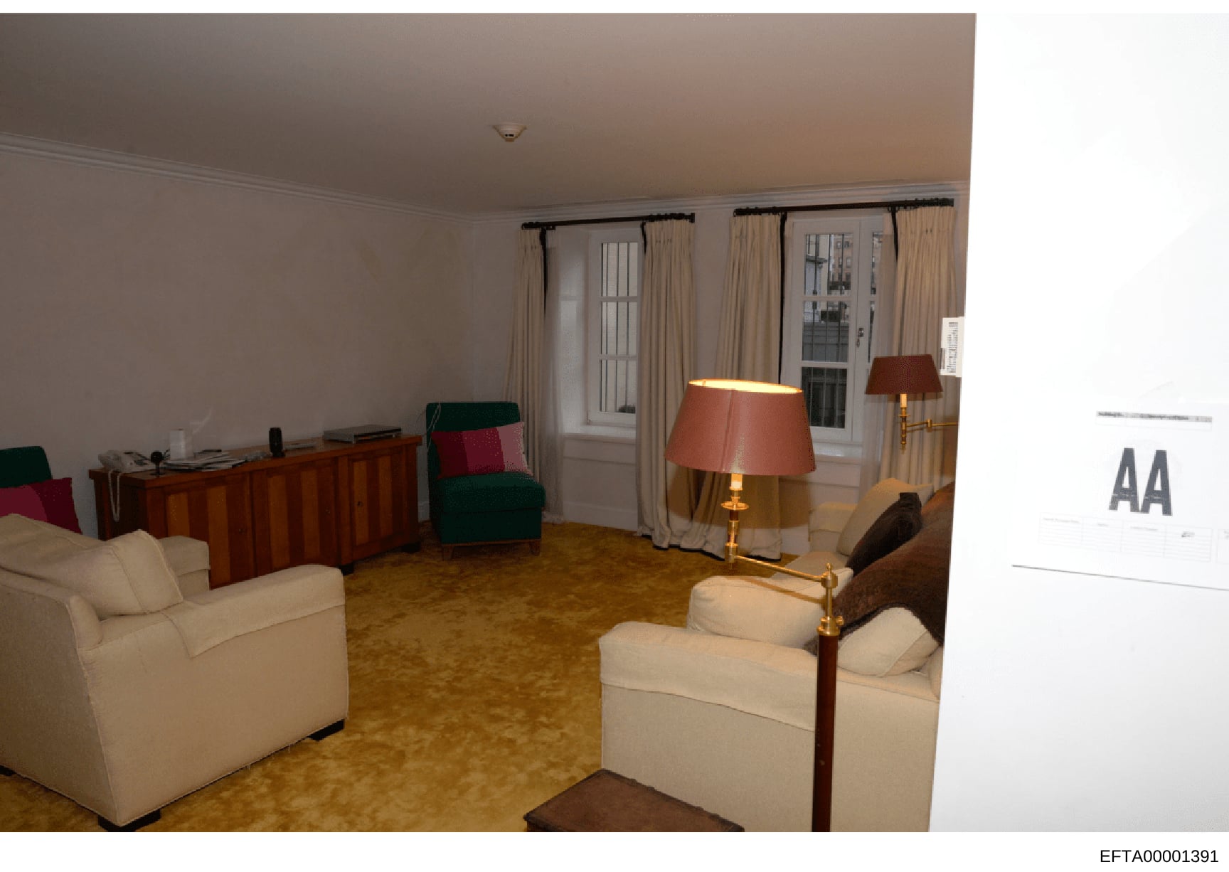 This is a photograph of an elegantly furnished residential living room featuring cream-colored upholstered furniture, wooden cabinetry, brass table lamps, and large windows with cream curtains. The room appears to be part of a high-end residence, lik