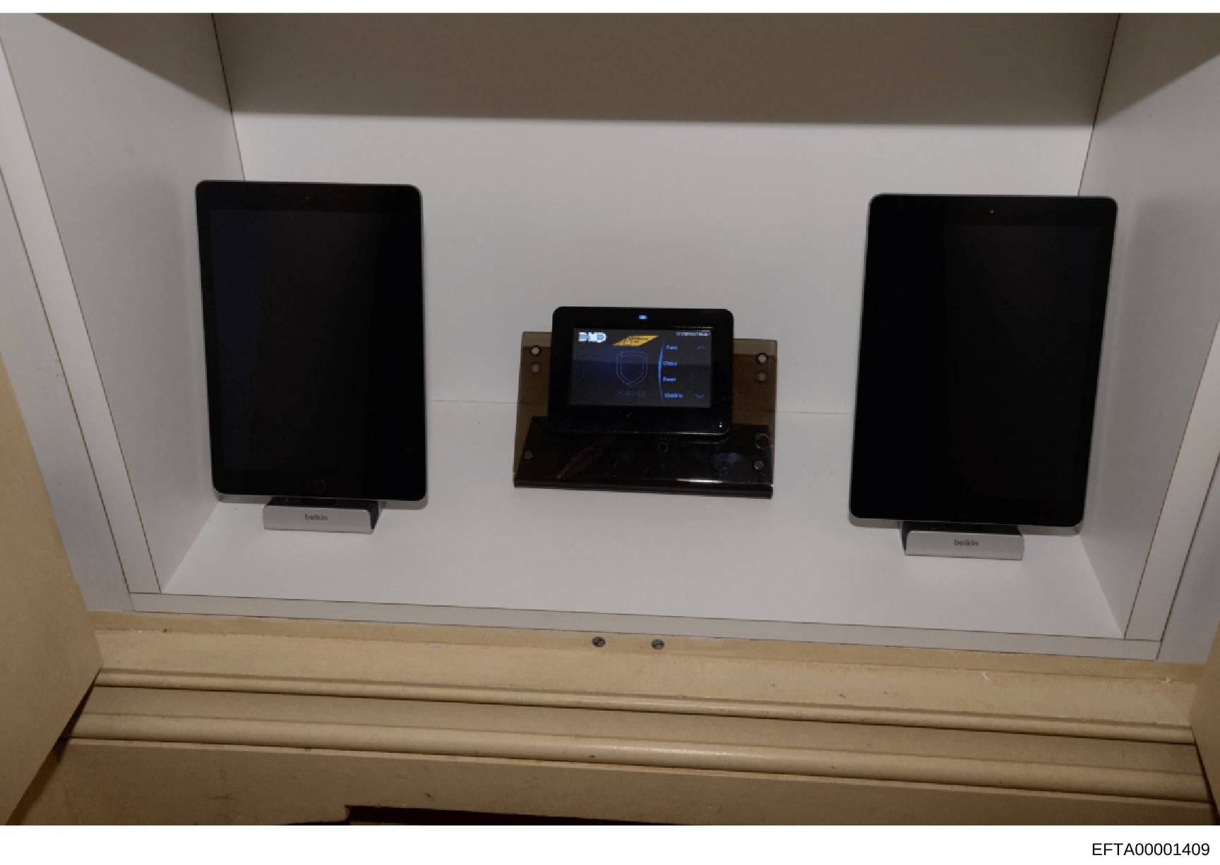 This photograph shows an interior wall-mounted display system consisting of three screens: two larger tablet-style devices on either side (marked 'belkin') and a central control panel with an illuminated interface displaying what appears to be a navi