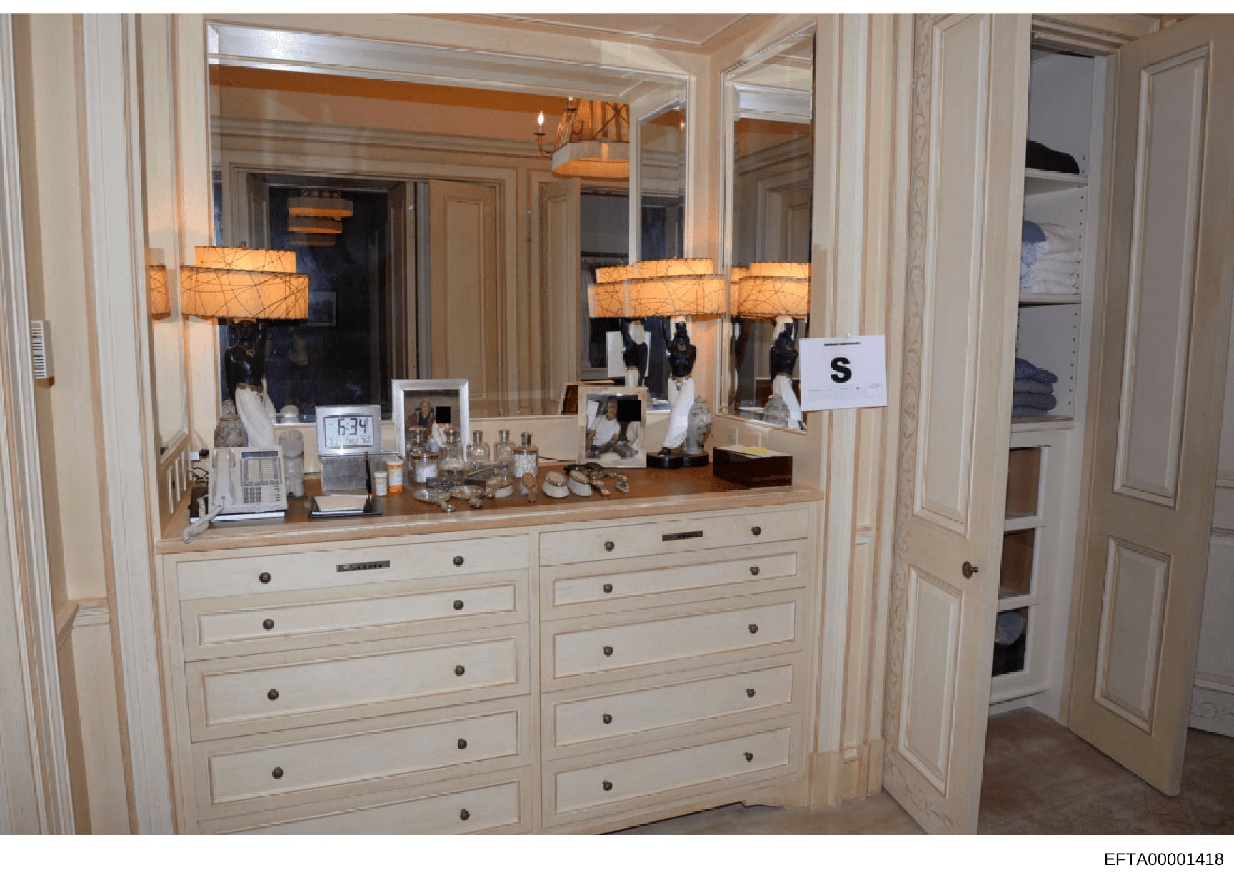 This photograph documents a high-end residential dressing area or master bedroom vanity, featuring custom cabinetry, multiple mirrors, decorative lighting, and personal items including framed photographs and grooming products. The image appears to be