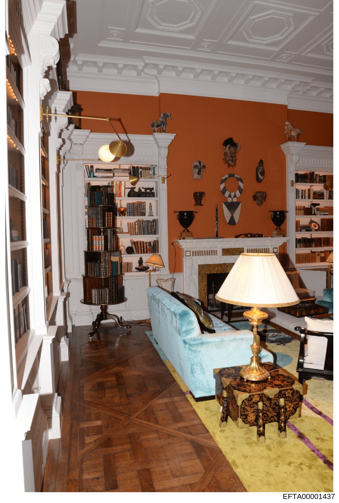 This photograph depicts an upscale interior room featuring classical architectural elements, extensive bookshelves, and decorative furnishings. The space includes built-in cabinetry, African tribal artwork and sculptures displayed on rust-colored wal